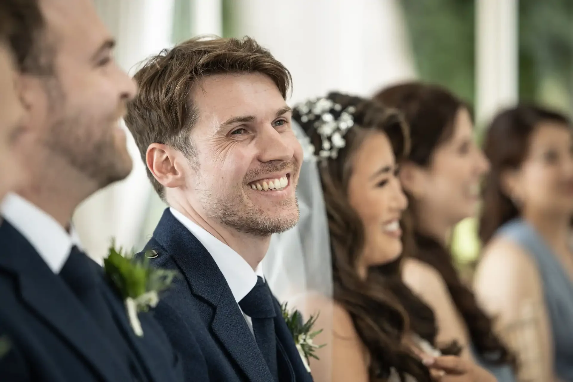 A man in a suit smiles while attending a formal event, likely a wedding. People in the background also appear cheerful, seated in a line.