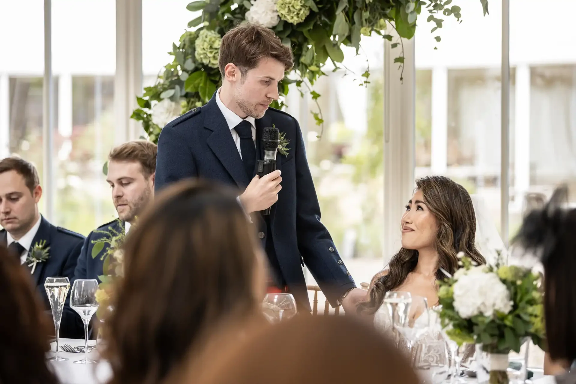 A man in a suit stands and speaks into a microphone at a formal event, while a woman seated beside him looks up and listens.