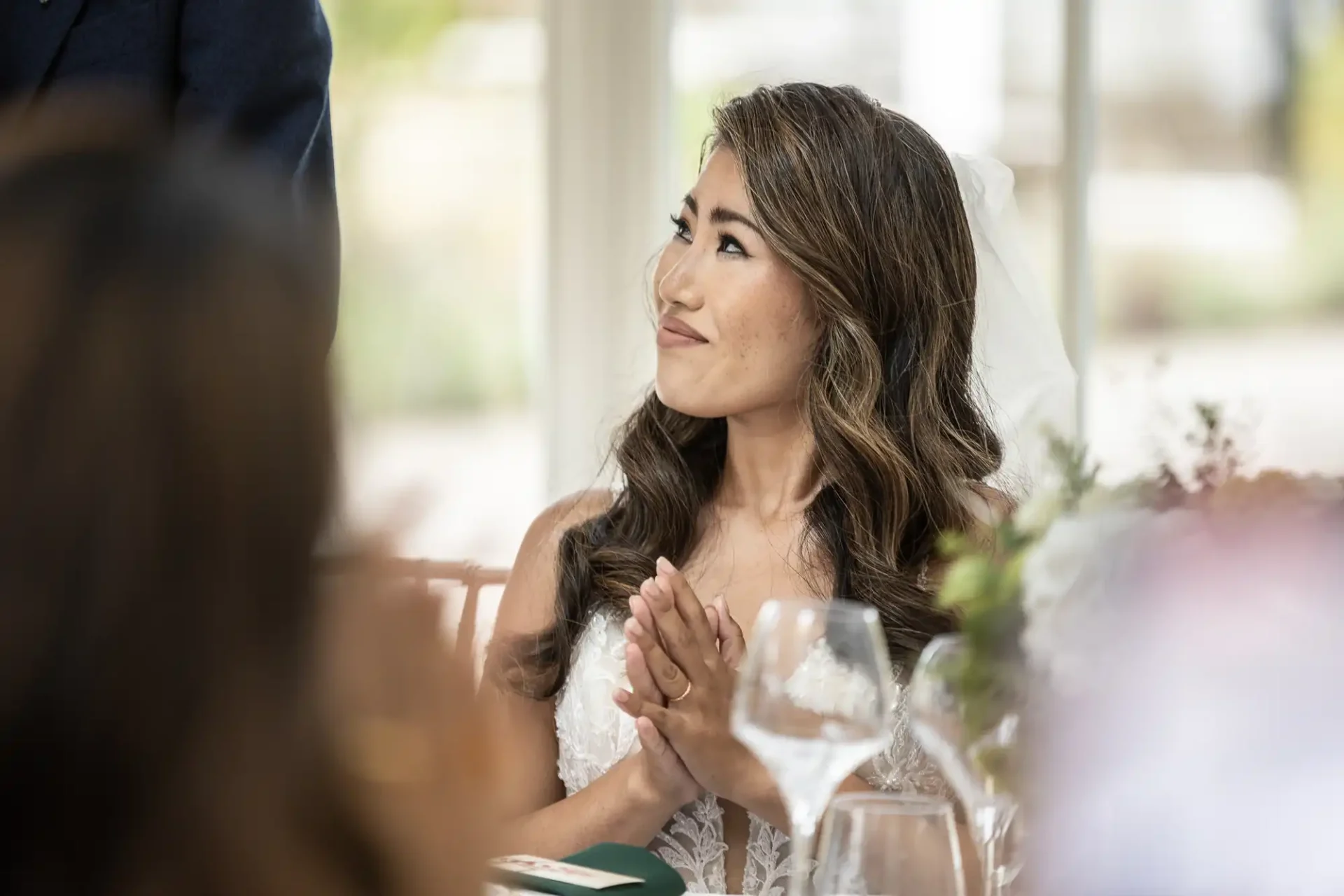 A woman in a white dress and veil sits at a table, clapping her hands and looking upward. There are glasses and a blurred person in the foreground.