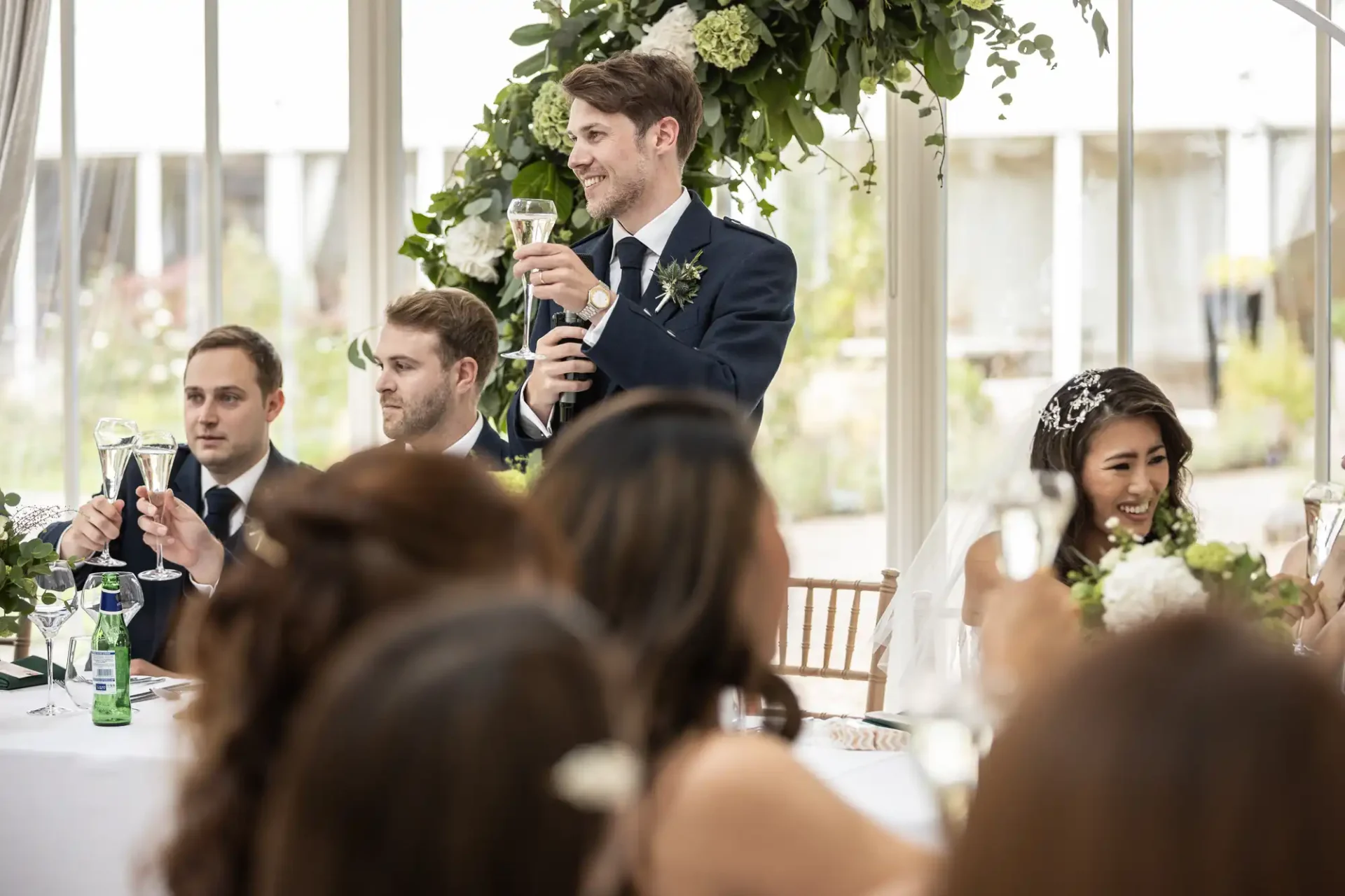 A man in a suit makes a toast at a decorated table during a celebration. Other people seated beside him hold champagne glasses, and one woman is wearing a veil.