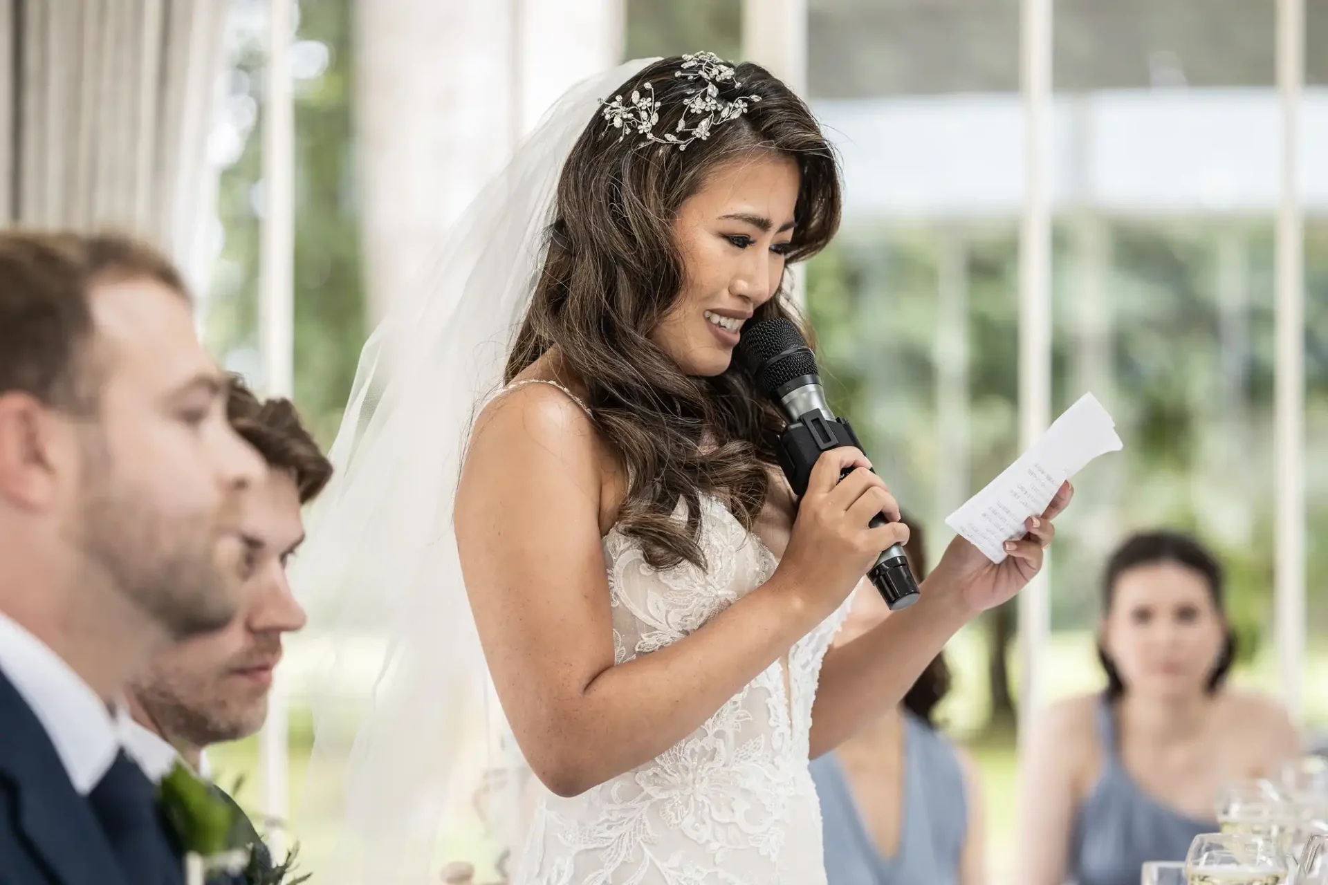 A bride in a white dress and veil reads from a paper while holding a microphone during a wedding speech. People are seated around her at a table.