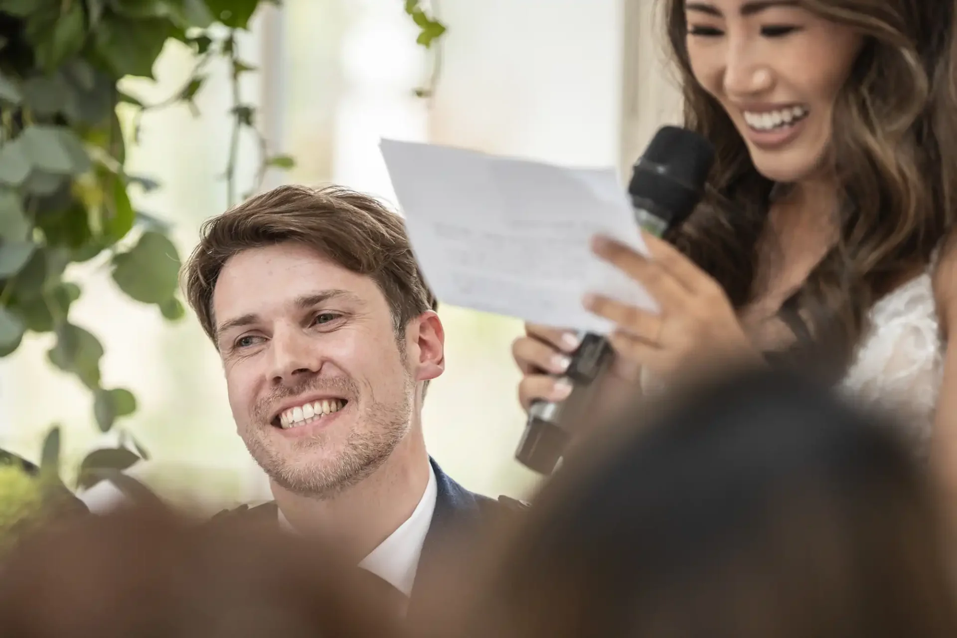 A woman holding a microphone and paper speaks, while a man seated beside her smiles, in a setting with greenery.