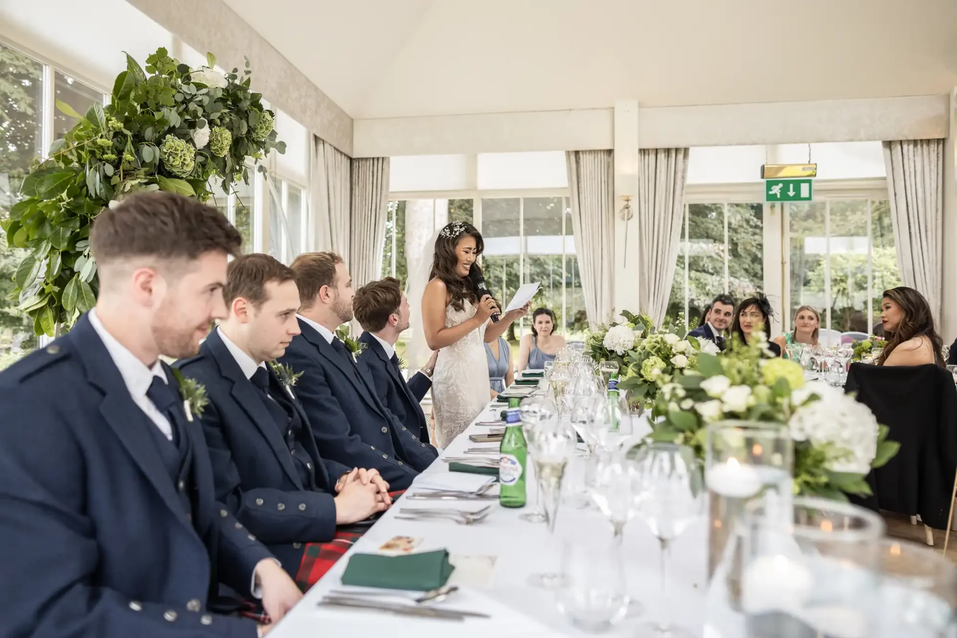 A woman stands and speaks at a wedding reception. She is in front of a table with people seated, including several men in kilts. The room is decorated with flowers.