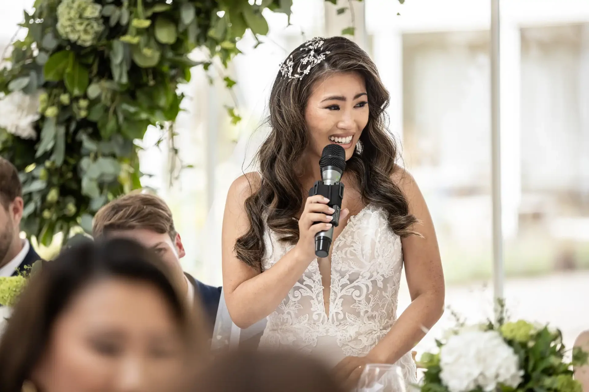 A bride in a white lace dress smiles while holding a microphone, standing under a floral arch at a wedding reception.