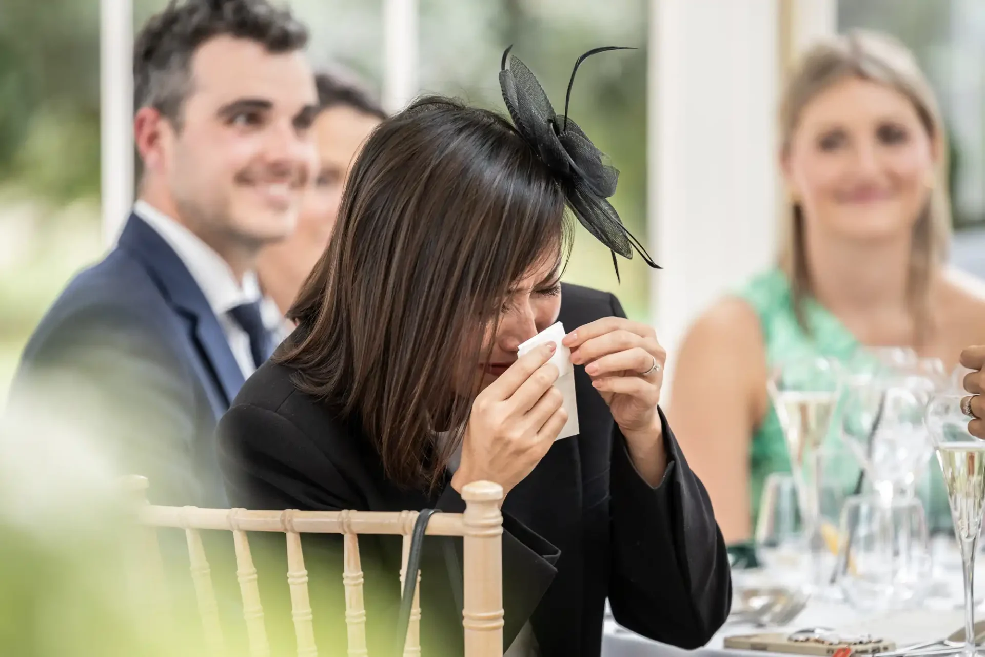 A woman in a black outfit and fascinator wipes her eyes with a tissue at a formal event, seated among other attendees.