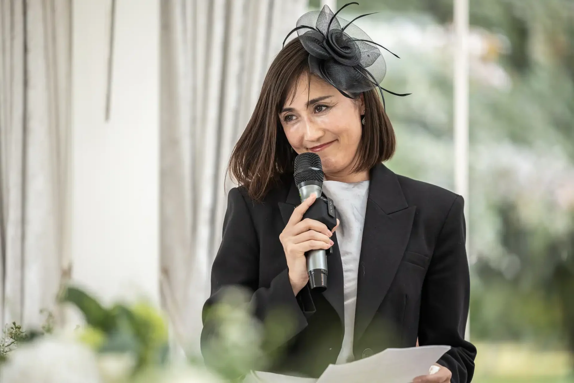 A woman in a black blazer and fascinator holds a microphone and a sheet of paper, standing indoors with a blurred background.