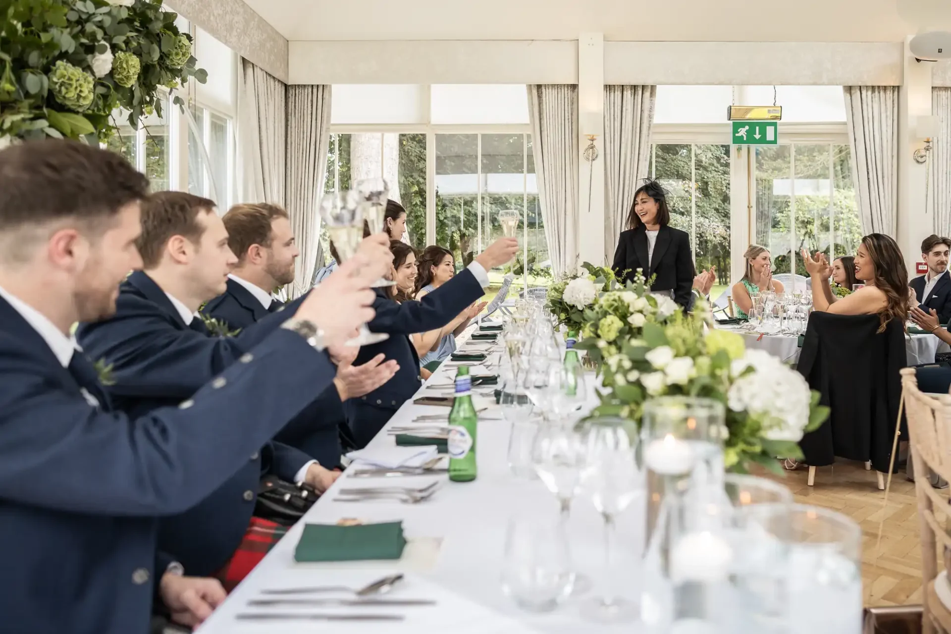 Guests raise glasses for a toast at a long, elegantly decorated table during a formal event in a bright room with greenery and large windows.