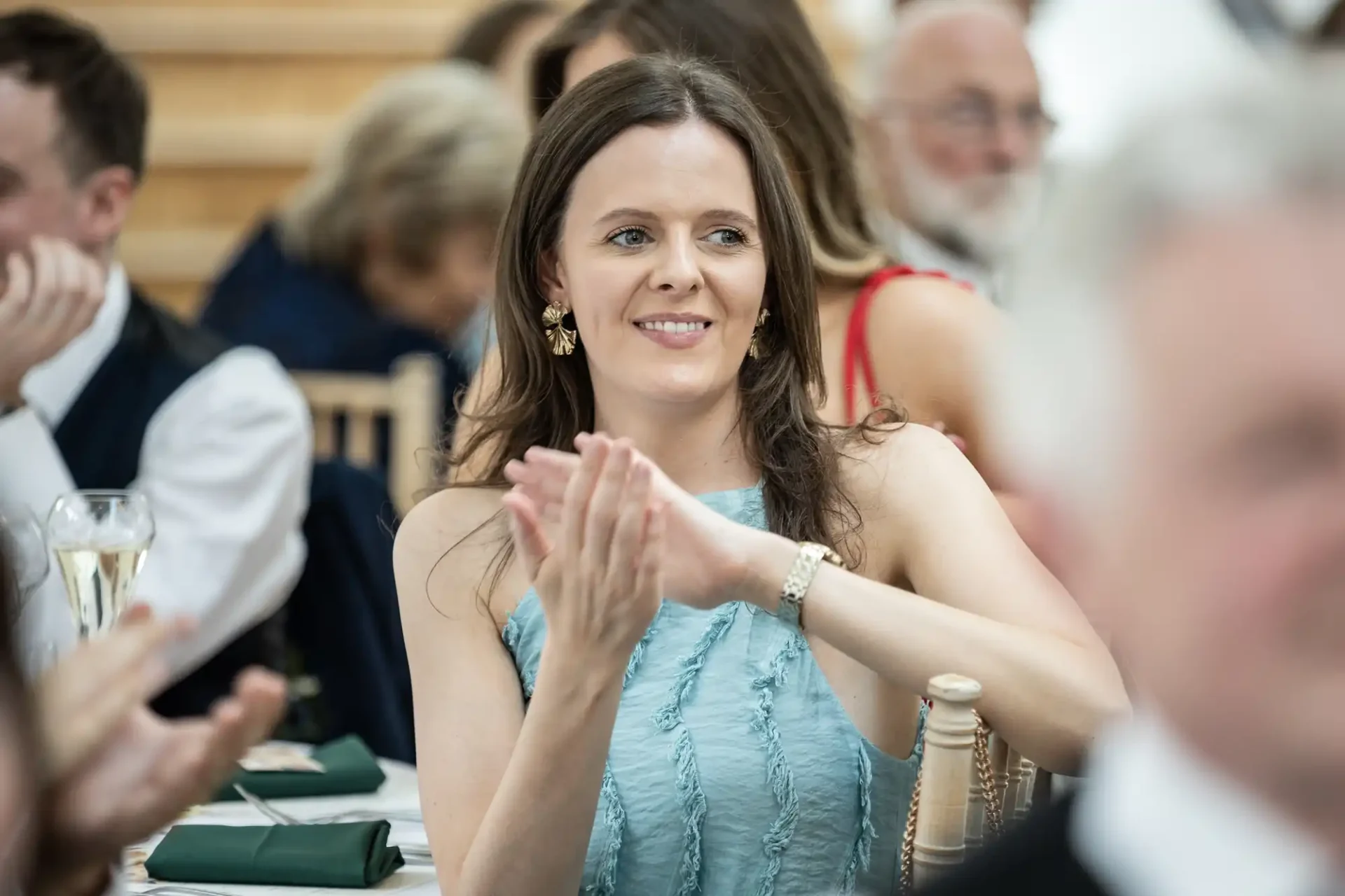 A woman in a light blue sleeveless top claps at a formal event, surrounded by seated people and tables with green napkins.