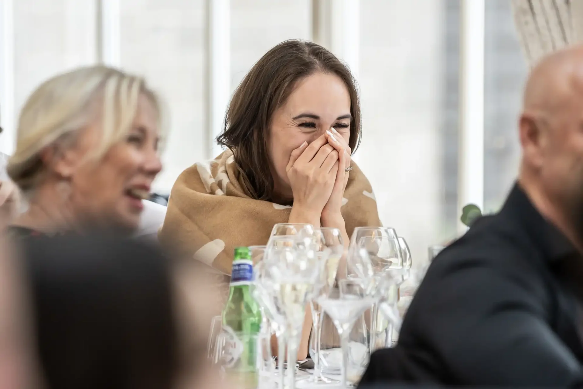 A woman seated at a dining table covers her mouth while laughing. She is surrounded by others, with empty wine glasses and a water bottle on the table.