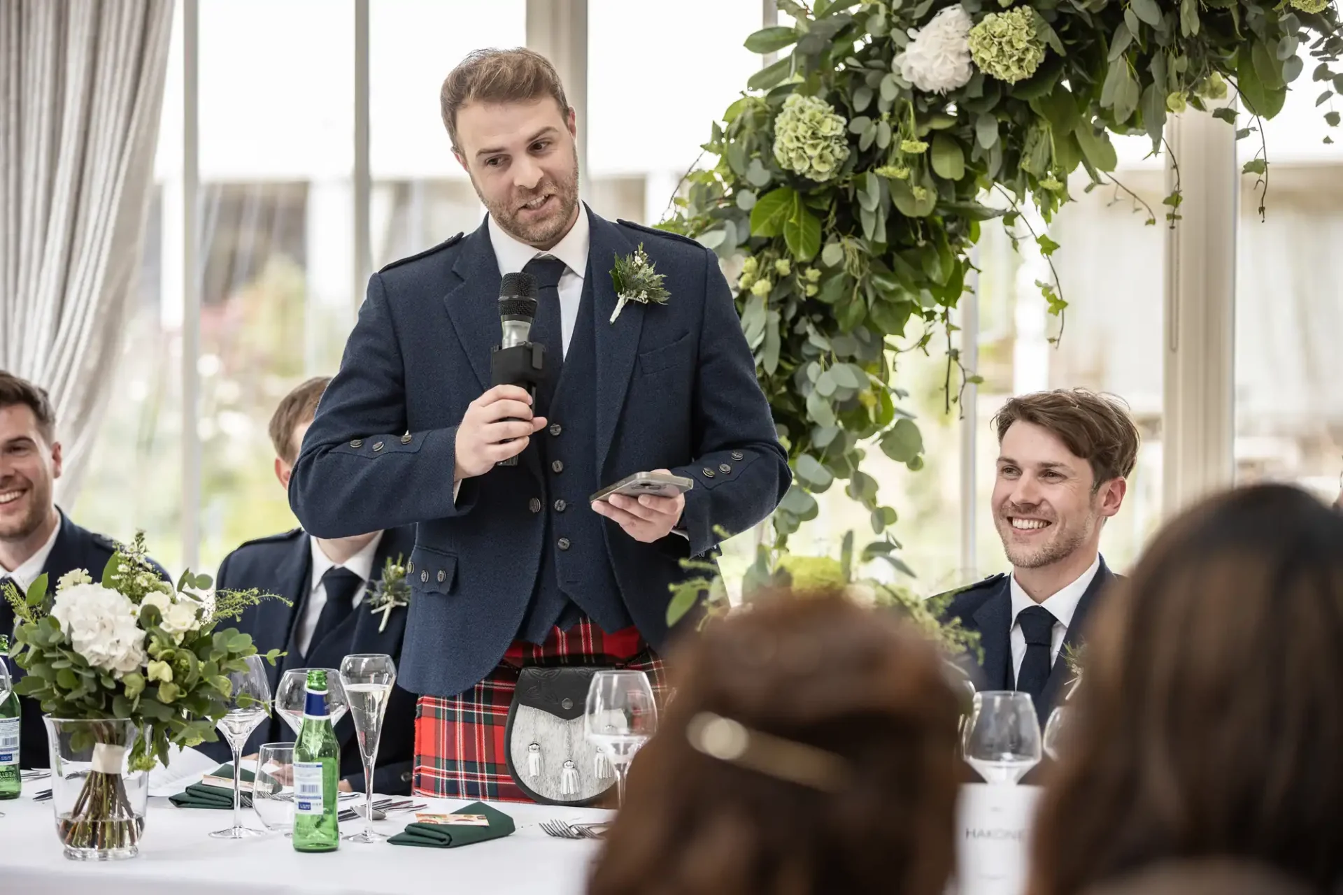 A man in a kilt gives a speech at a wedding reception table with guests. Floral decorations and table settings are visible.