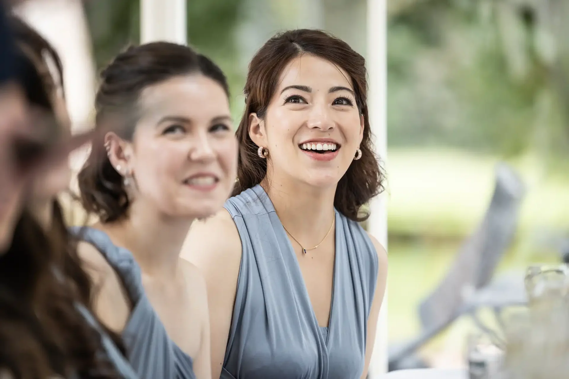 Two women in sleeveless blue dresses sit at a table outdoors, smiling and looking in the same direction.