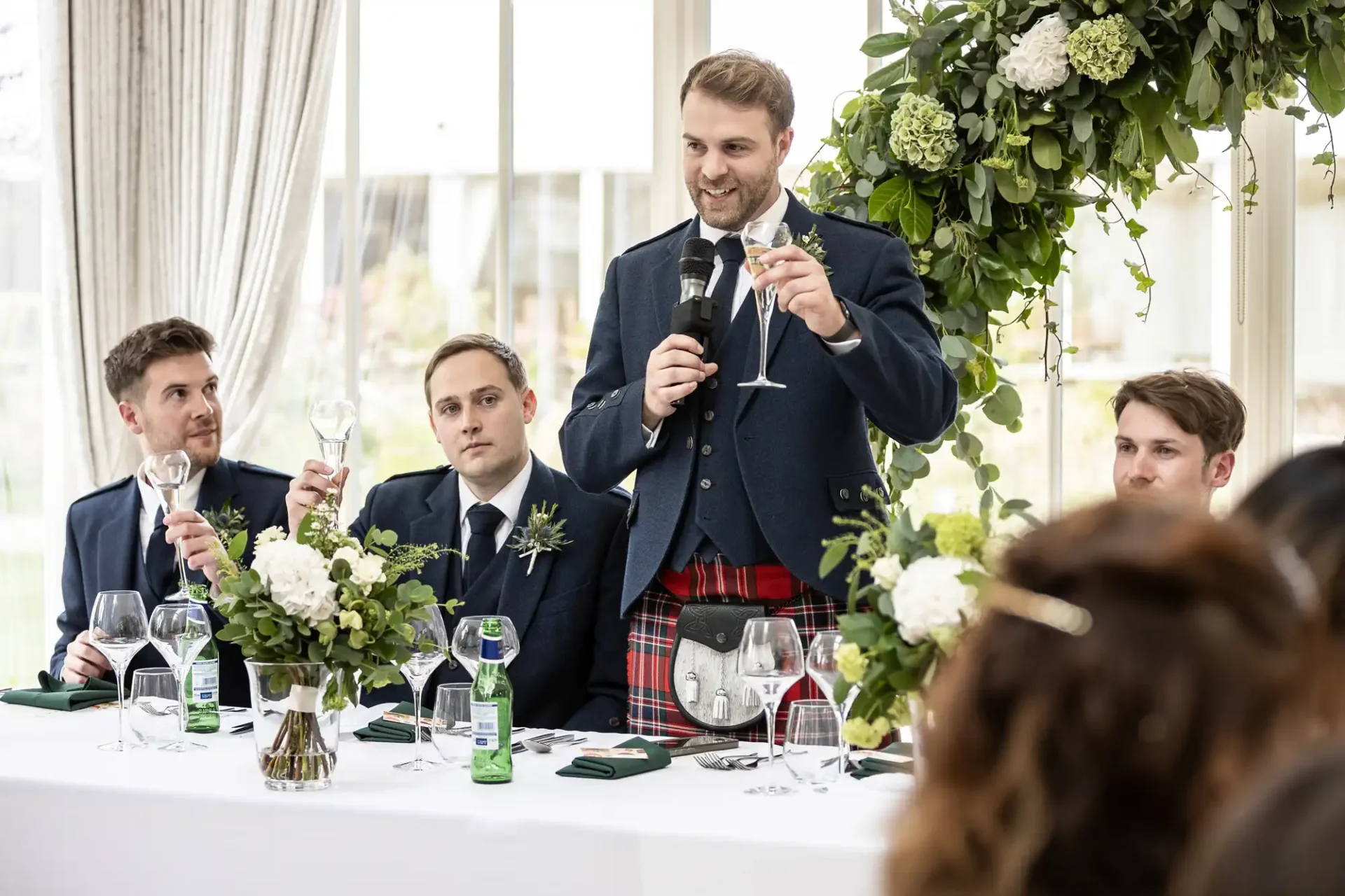 A man in a kilt gives a speech at a wedding reception, holding a microphone and a glass. Three seated men in suits listen attentively. Floral decorations and tableware are visible.