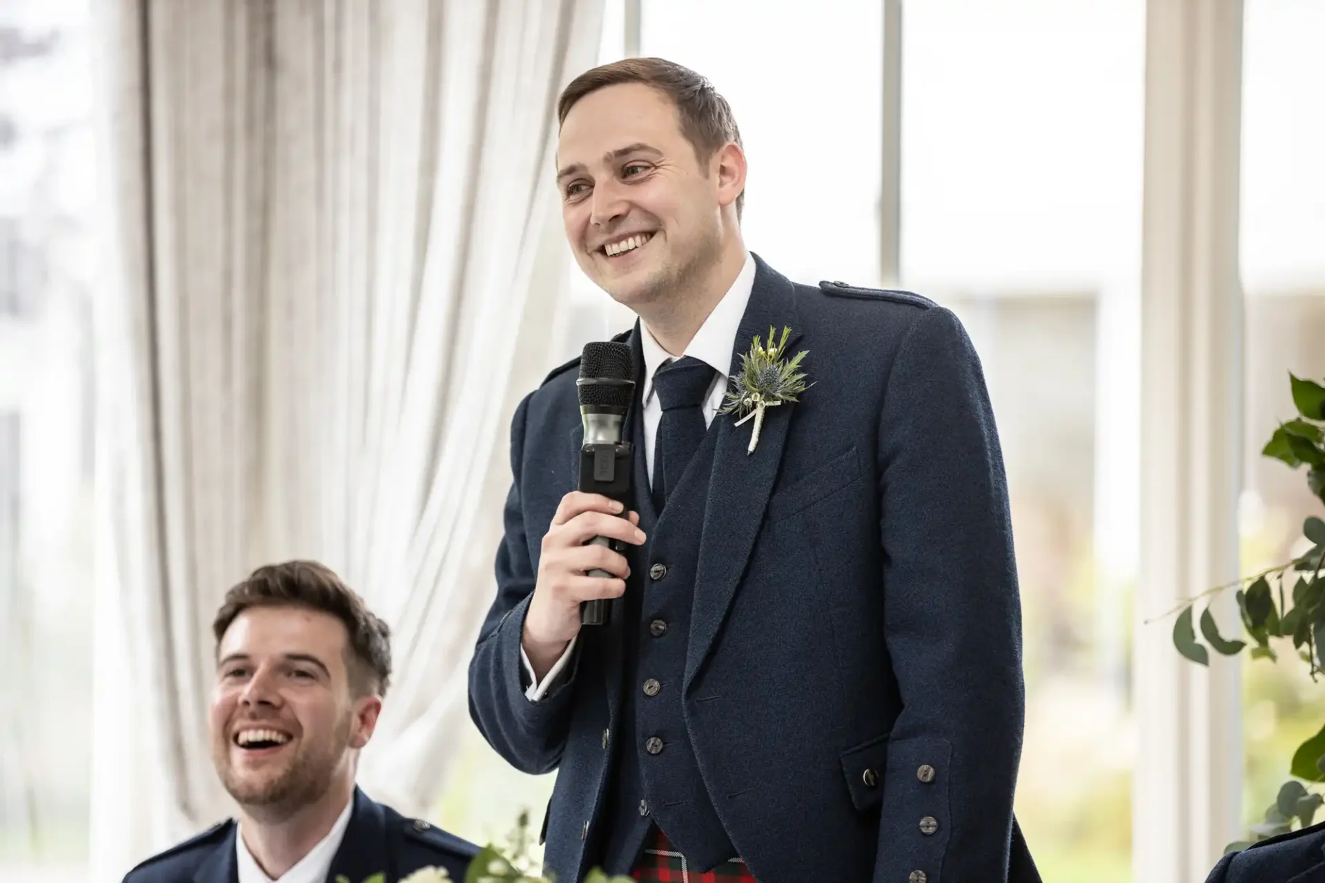 Man in formal attire speaking into a microphone at a gathering, with another seated man smiling in the background.