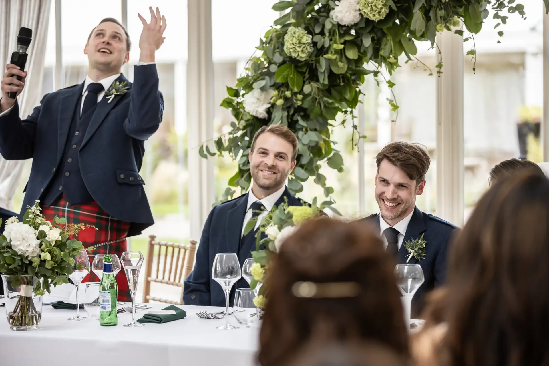 Man in kilt passionately speaking into microphone at a wedding reception; two men in suits smile while seated at a decorated table.