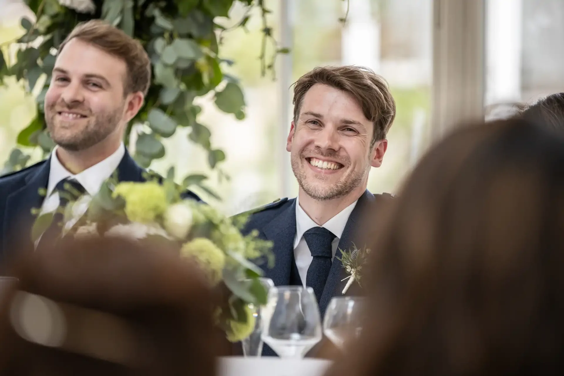 Two smiling men in suits sit at a table with floral arrangements, blurred foreground figures visible.