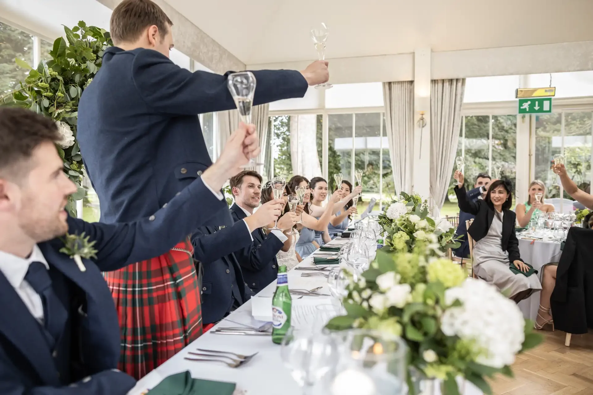 Wedding reception scene with guests raising glasses in a toast. The room has white walls, large windows, and floral decorations. A man in a kilt stands, leading the toast.