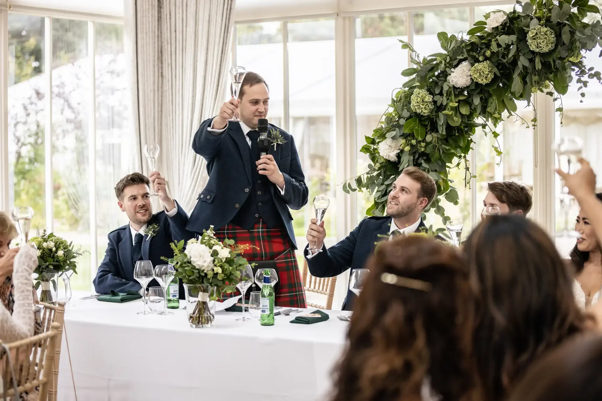 A man in a kilt makes a toast at a wedding reception, surrounded by seated guests raising glasses. A floral arrangement decorates the table.