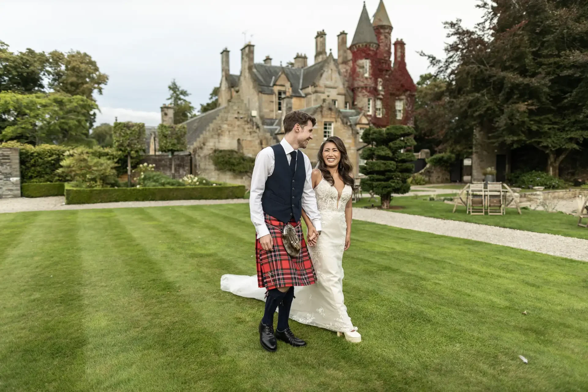 A bride and groom walk on a lawn in front of a castle-like building. The groom wears a kilt, while the bride is in a white gown. The background shows gardens and trees.