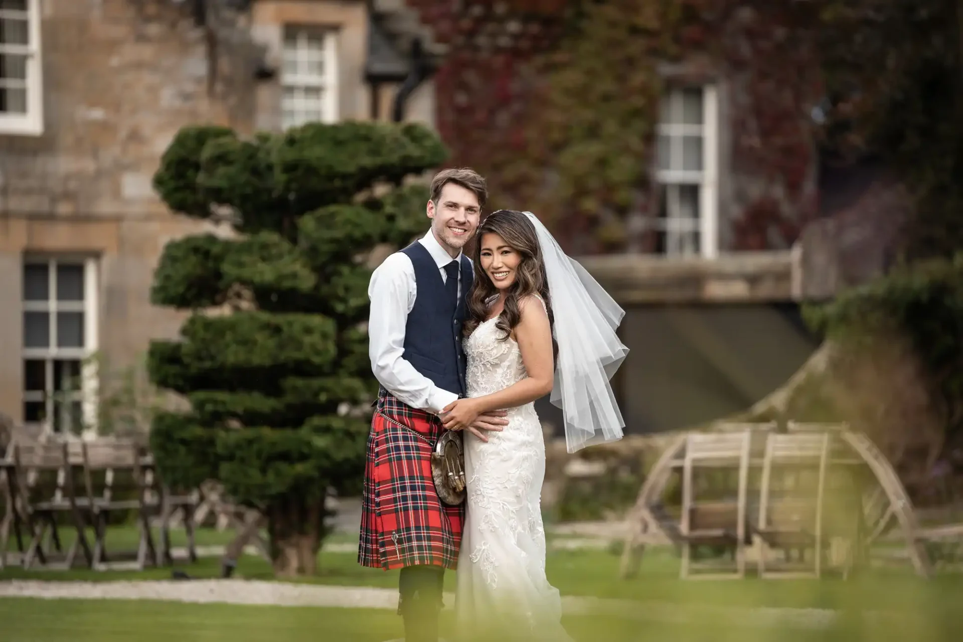 Bride and groom smiling, standing in front of a large tree and stone building. The groom is wearing a kilt, and the bride is in a white wedding dress with a veil.