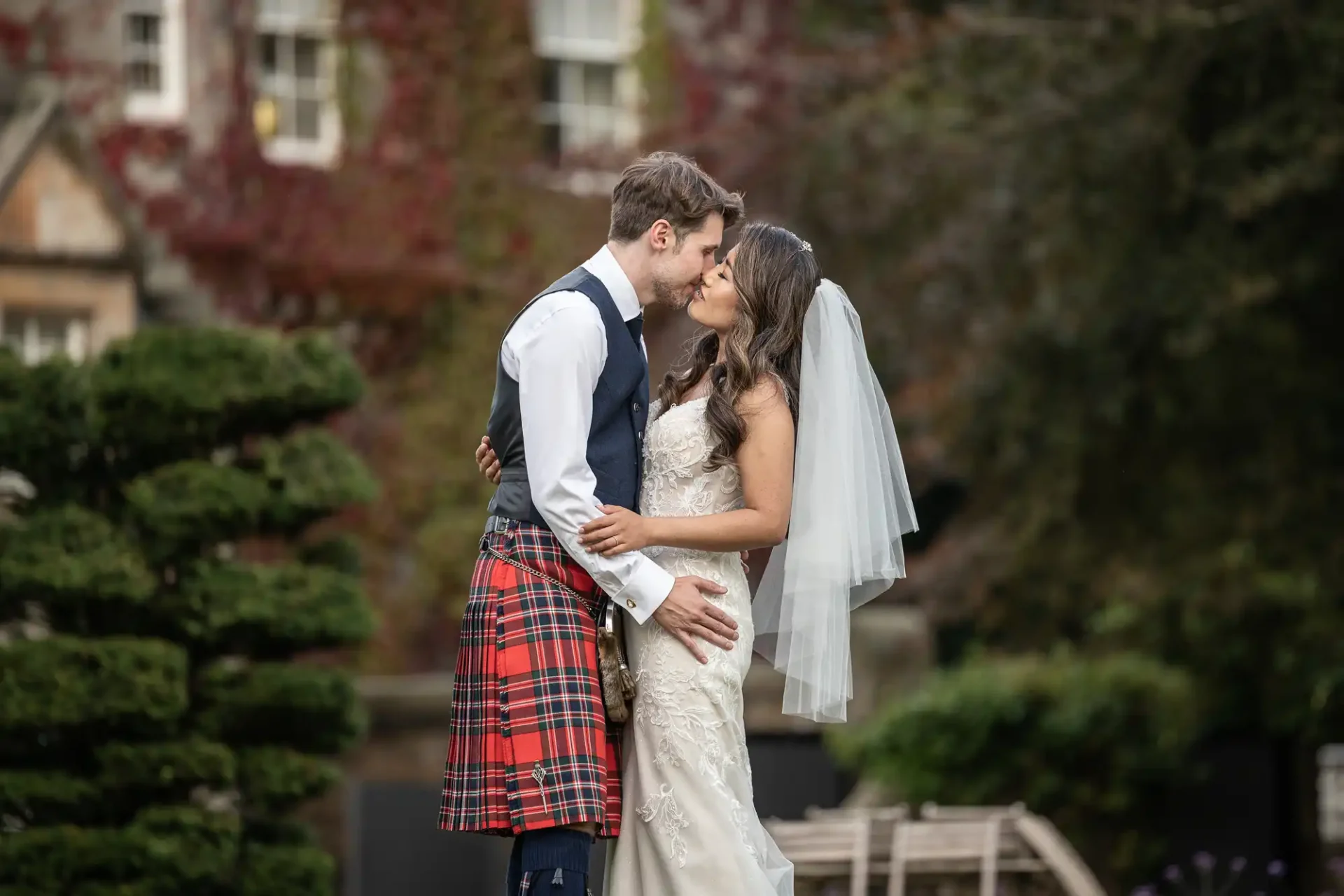 Bride in a white dress and groom in a tartan kilt are kissing outdoors in a garden setting.