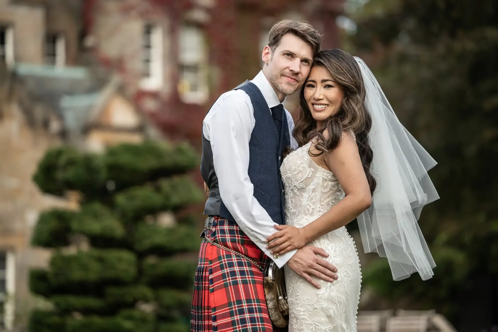 Carlowrie Castle wedding photographer: A bride in a white dress and veil stands next to a groom wearing a kilt and vest, both smiling, in front of a lush garden backdrop.