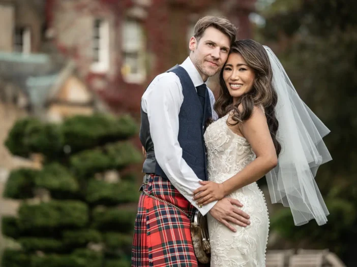 Carlowrie Castle wedding photographer: A bride in a white dress and veil stands next to a groom wearing a kilt and vest, both smiling, in front of a lush garden backdrop.