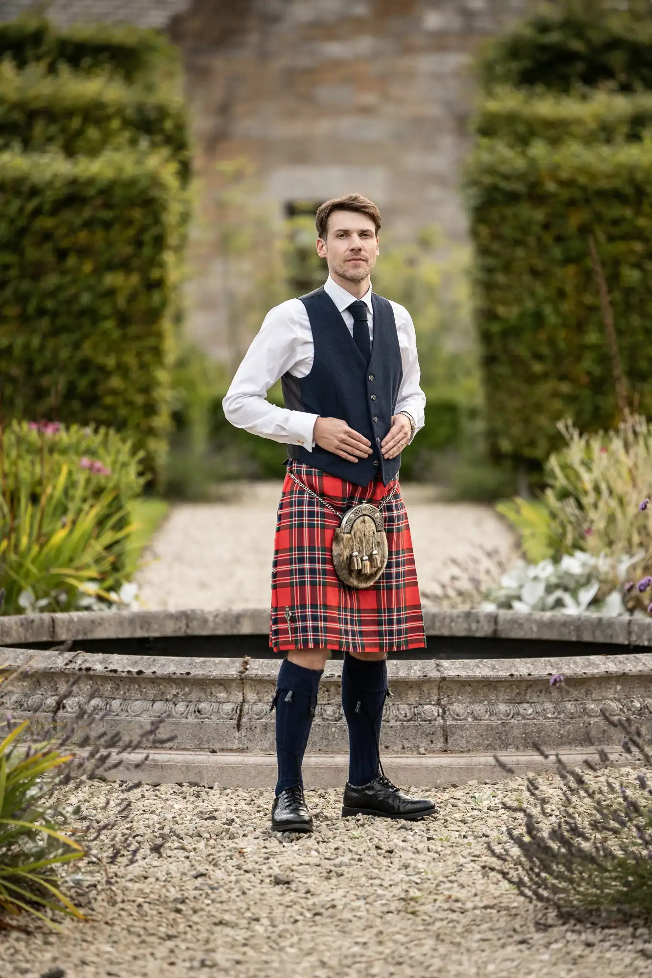 Man wearing a traditional Scottish kilt, vest, and tie stands outdoors on a gravel path in a garden setting.