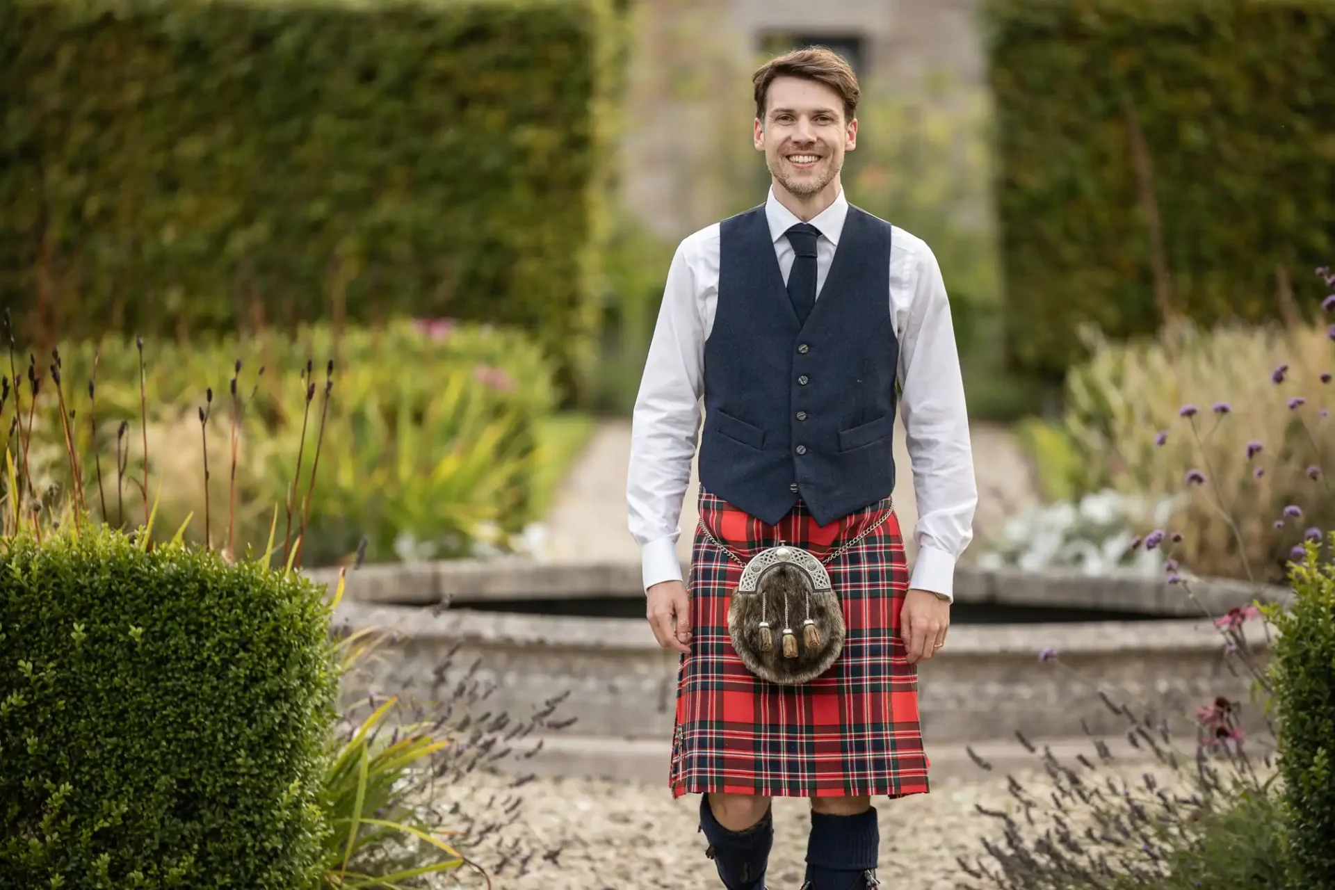 A man wearing a tartan kilt, vest, and tie stands in a garden with greenery and flowers.