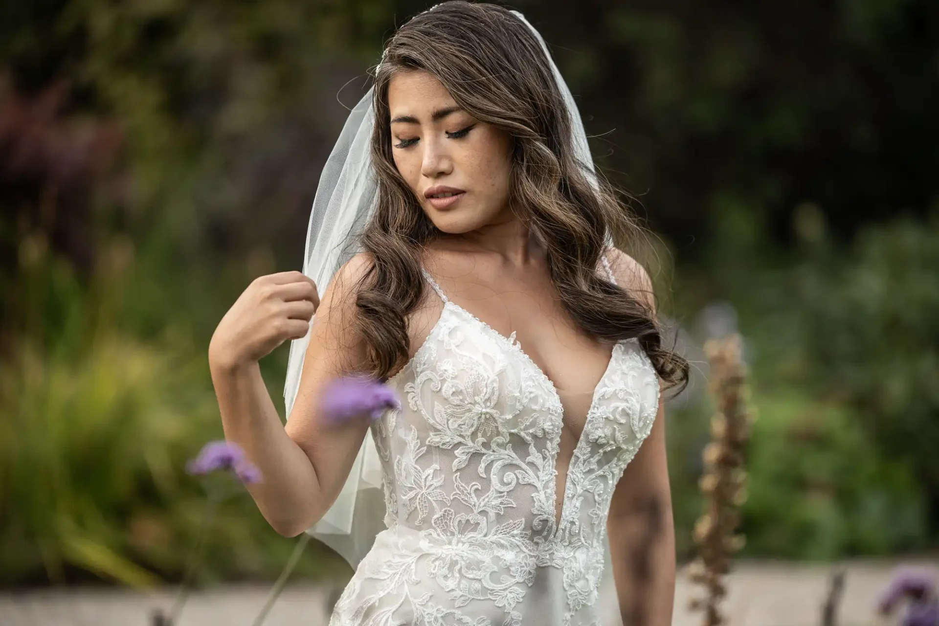 A woman in a white wedding dress and veil stands outdoors, surrounded by greenery and purple flowers.