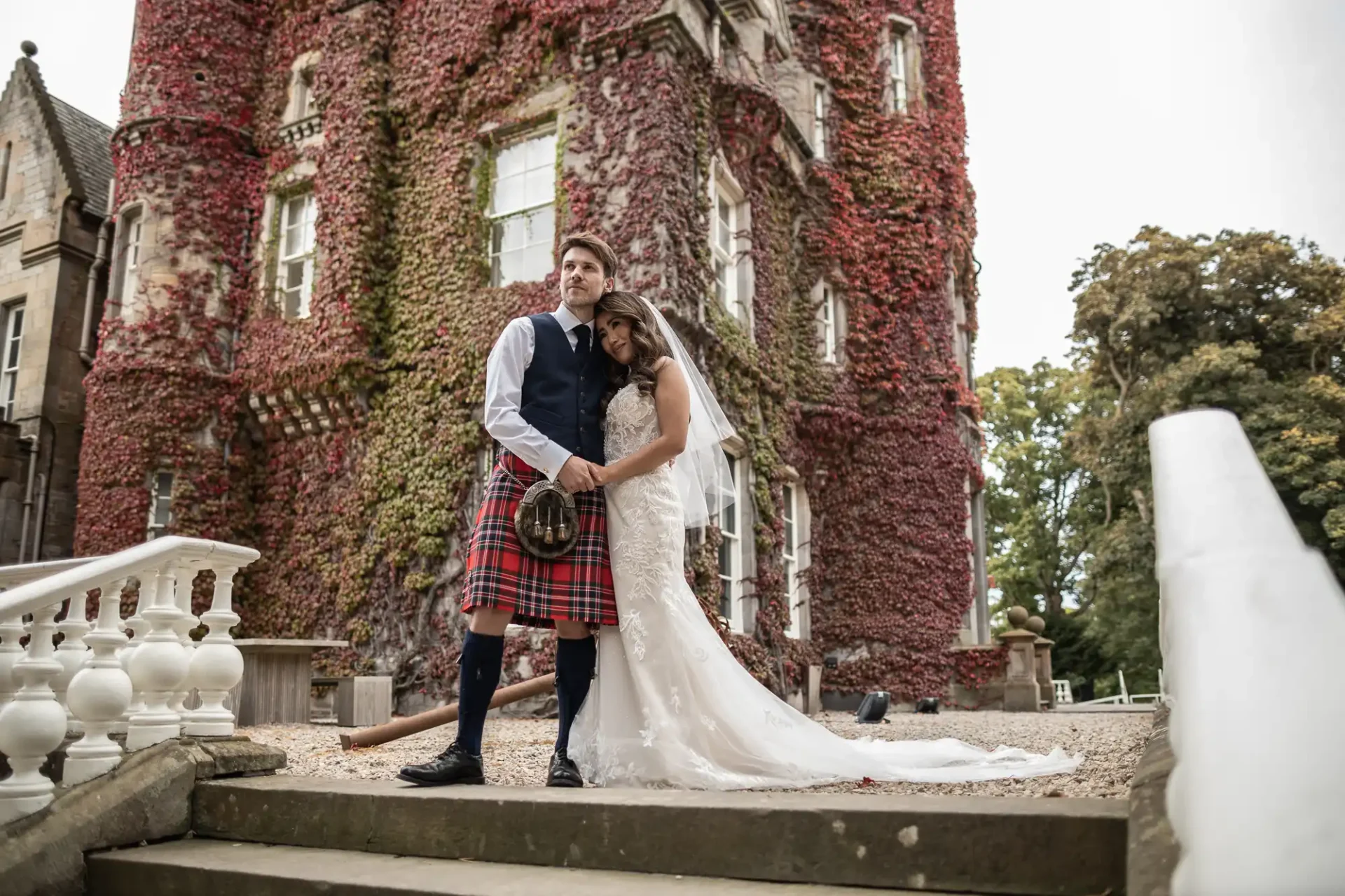 Bride in a wedding dress and groom in a kilt stand embraced in front of an ivy-covered, red-brick castle with white stairs in the foreground.