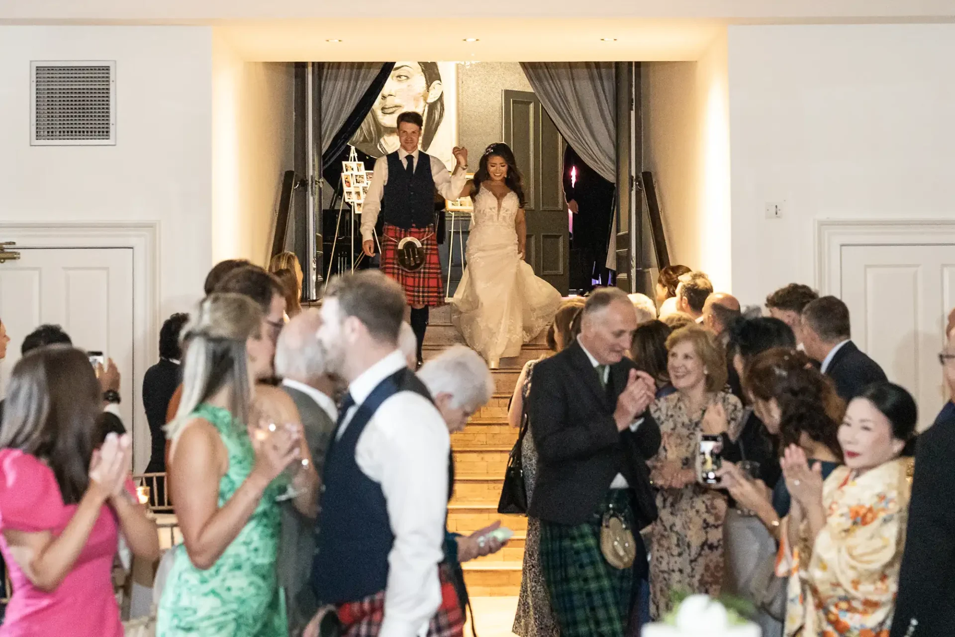 Bride and groom descend stairs at a wedding, surrounded by applauding guests. Some men wear kilts. A portrait is visible in the background.