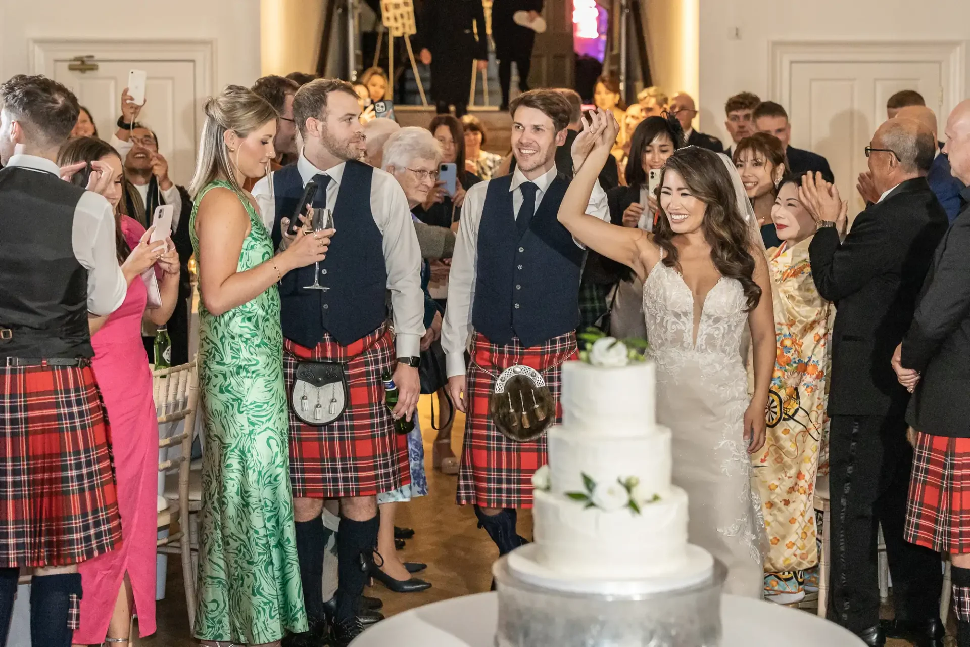 Bride and groom walk through cheering guests, some in kilts, at a wedding reception. A white tiered cake is in the foreground.