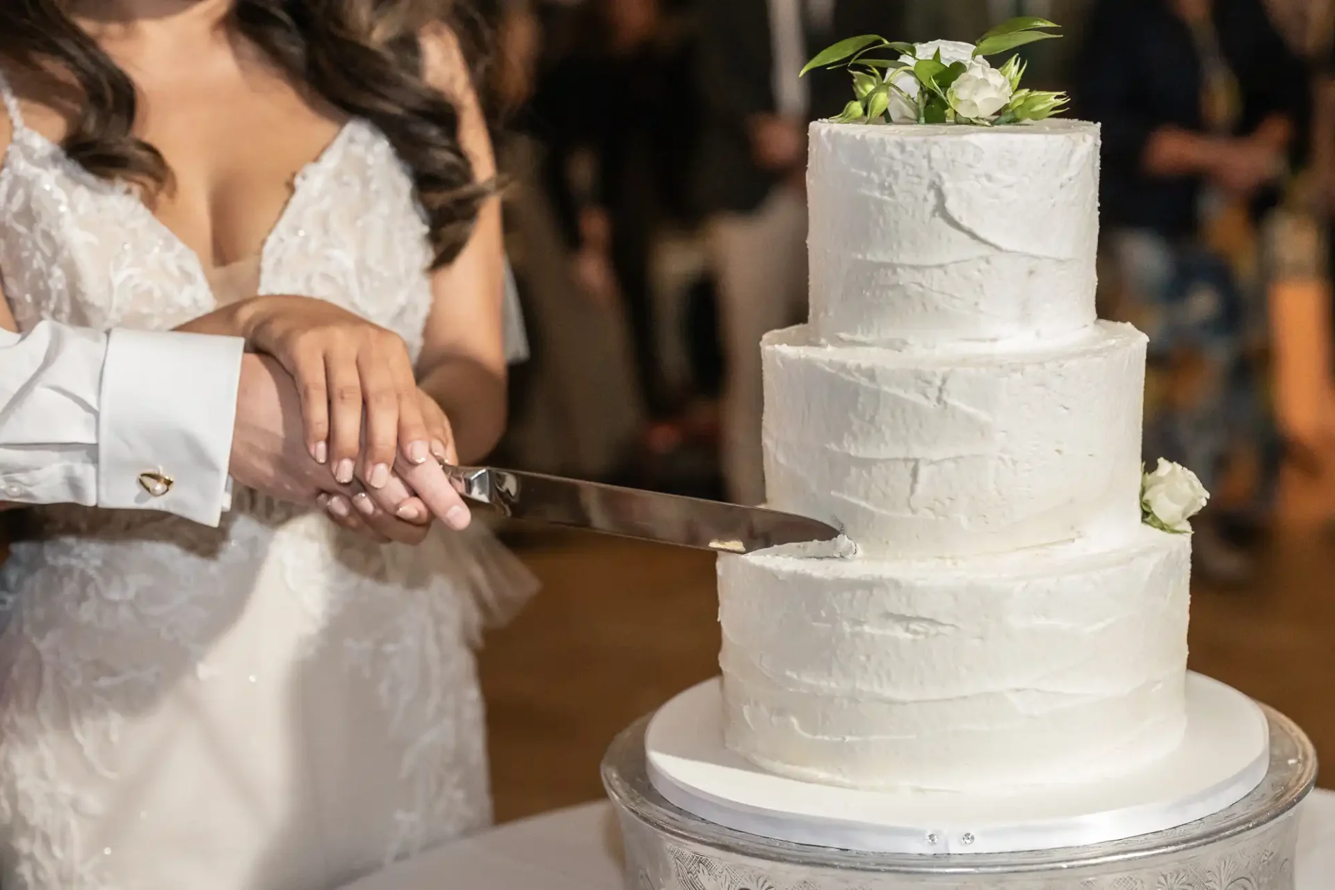 A couple cuts a three-tiered white wedding cake topped with flowers. The bride wears a white dress, and the groom's hand is in a white shirt, both holding the knife together.