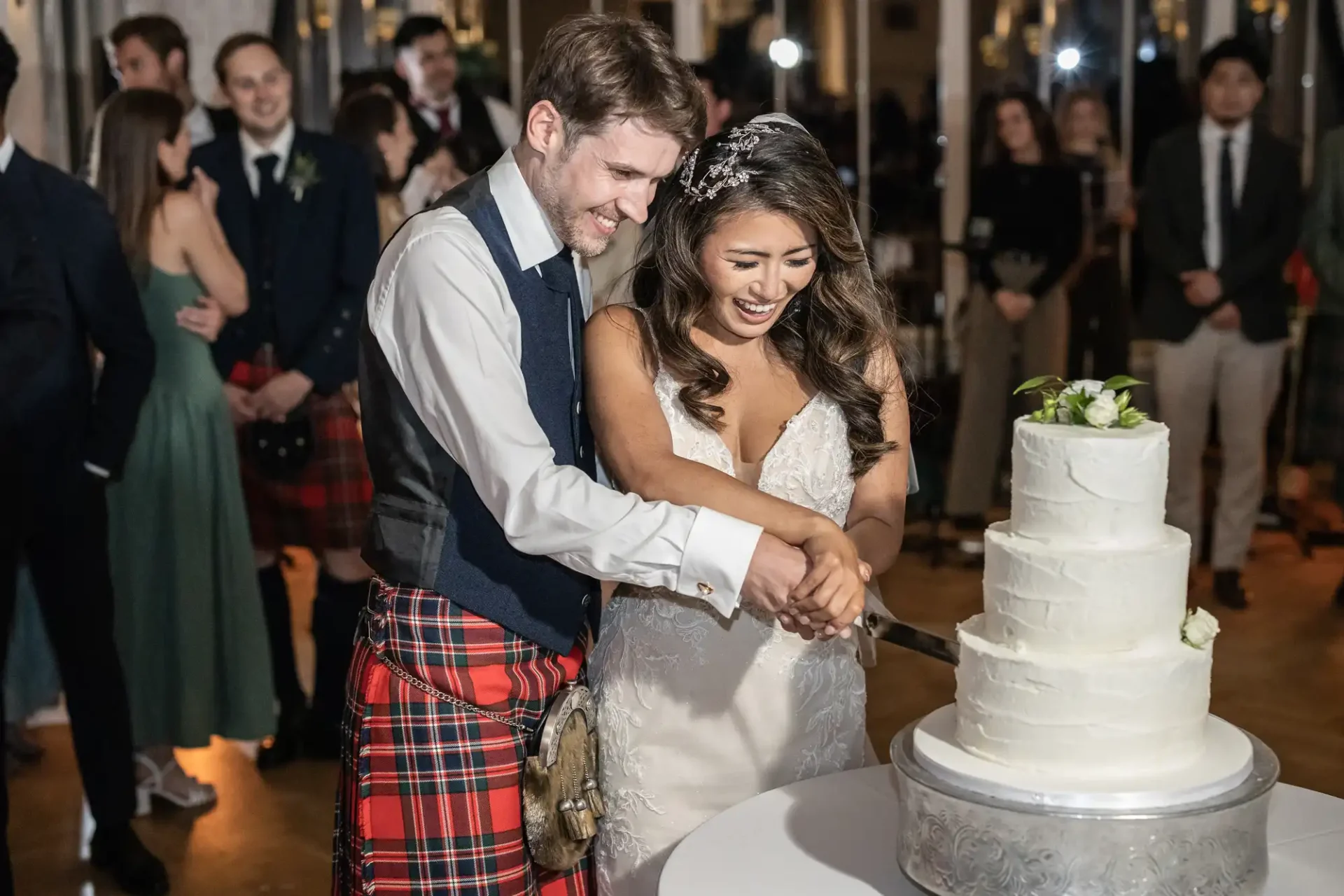 A bride and groom are cutting a white, multi-tiered wedding cake. The groom is wearing a kilt, and the bride is in a white dress. Guests are visible in the background.