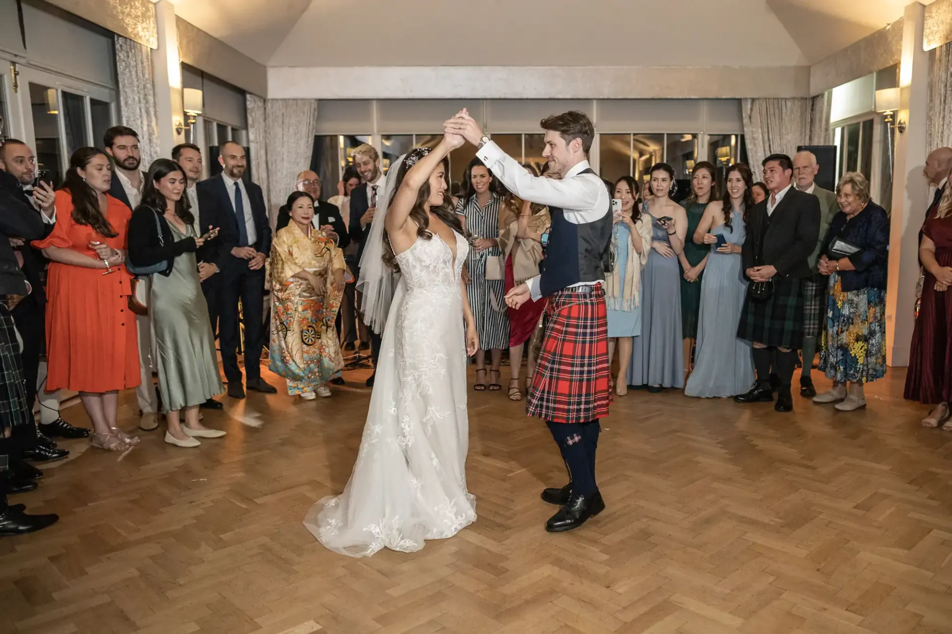 A bride and groom dance in the center of a room, surrounded by people watching. The groom wears a kilt, and the bride is in a white gown.