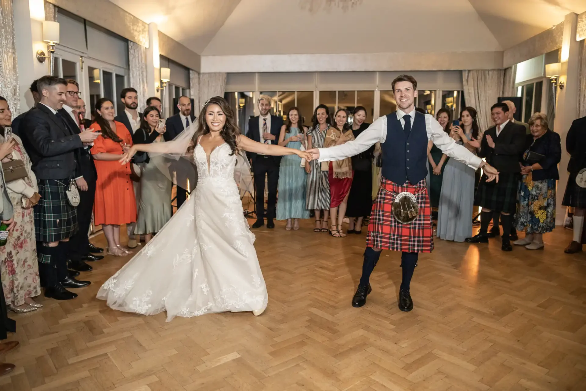 Bride and groom hold hands while dancing in front of guests at a wedding reception. The groom wears a kilt, and the bride is in a white gown.