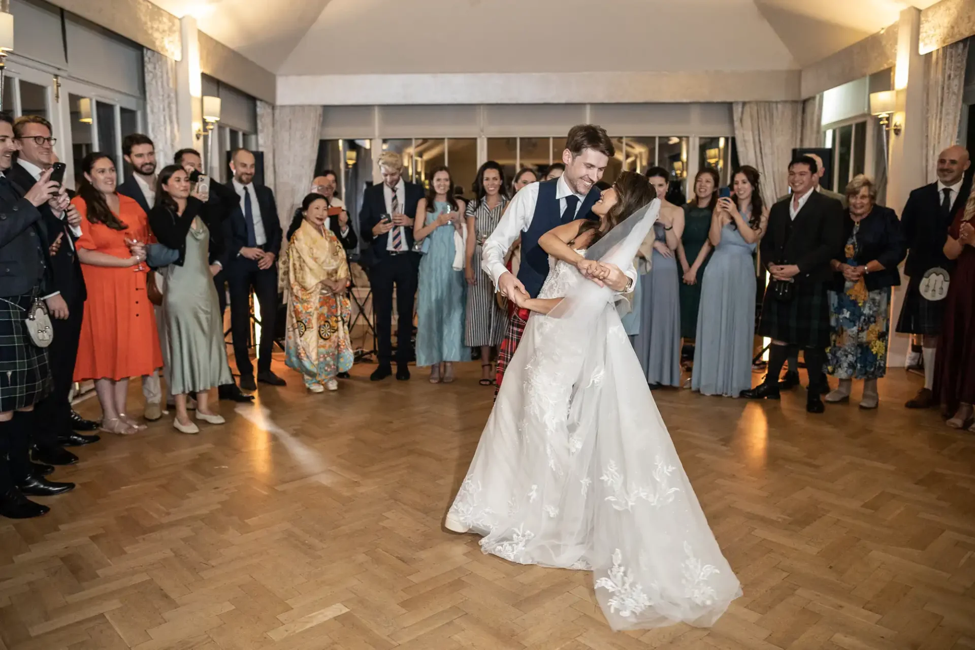 A bride and groom share their first dance in a large room. Guests stand around them, watching and smiling.