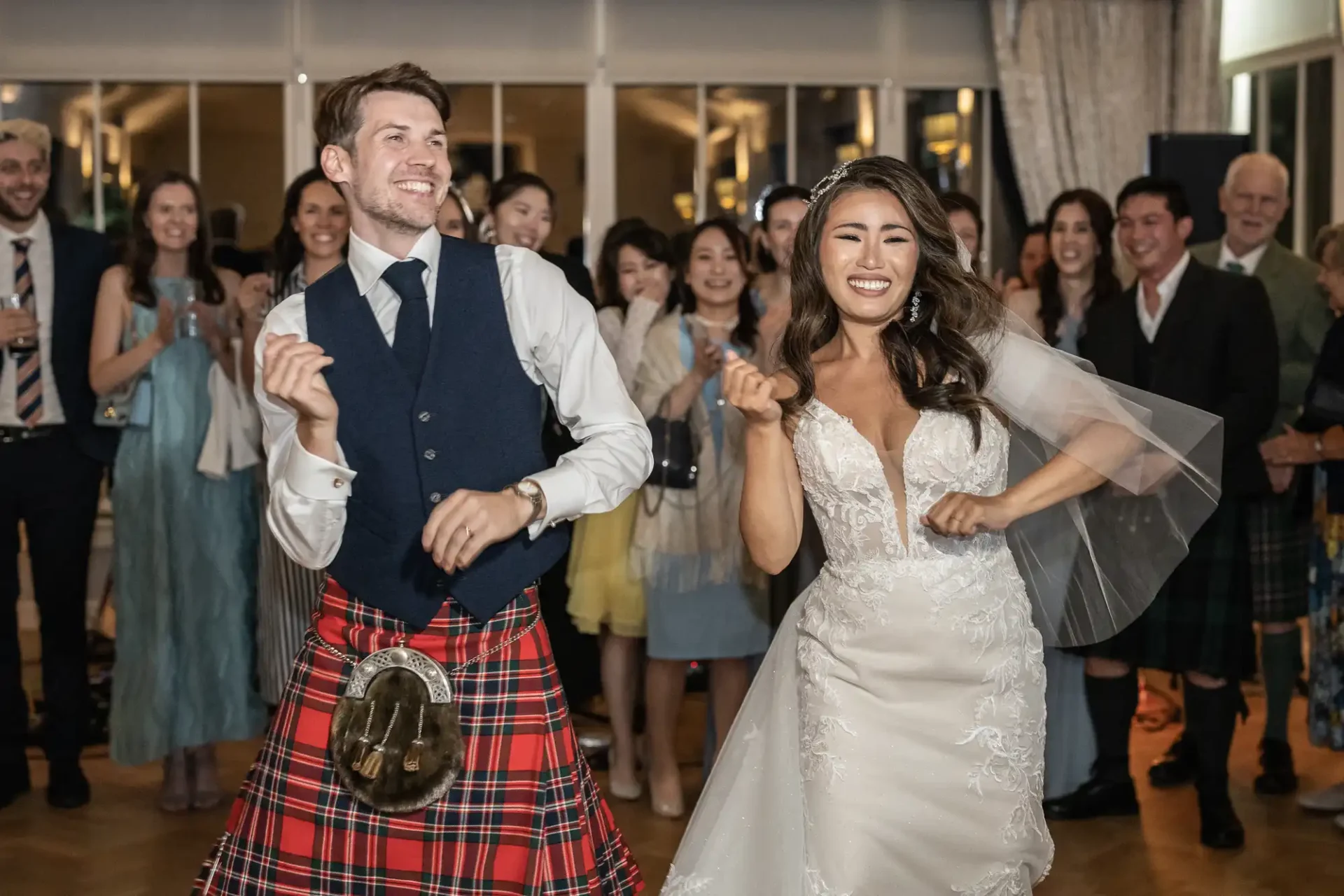 A bride and groom are dancing joyfully at their wedding, surrounded by smiling guests. The groom is wearing a kilt, and the bride is in a white gown.