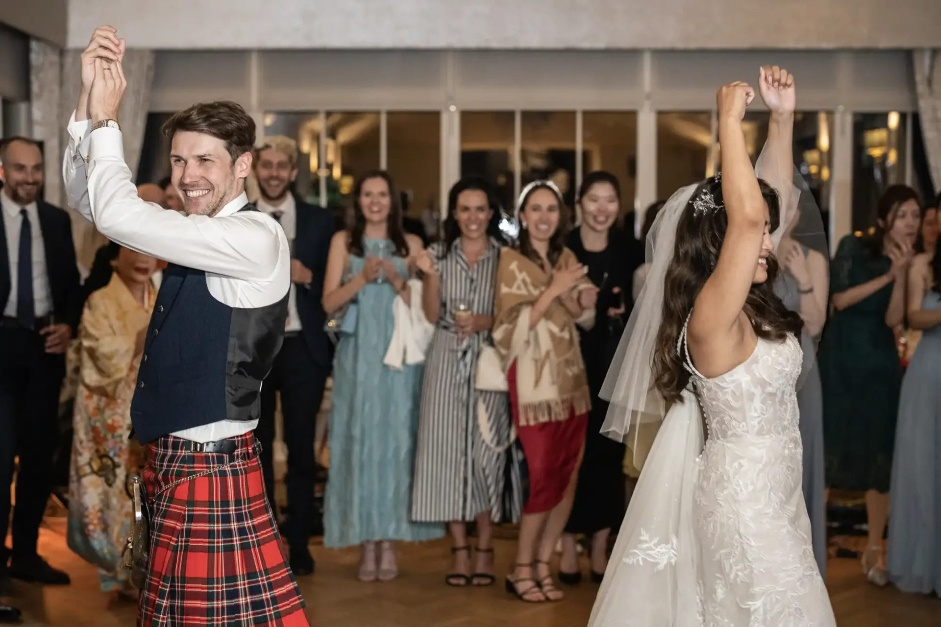 A bride and groom celebrate on the dance floor, surrounded by smiling guests. The groom wears a kilt, and the bride is in a white dress.