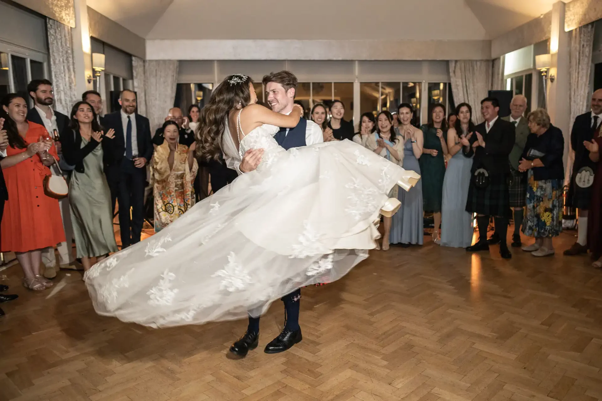 A couple dances in a large room, the groom lifting the bride, surrounded by smiling guests. The bride's gown flares out as they move.