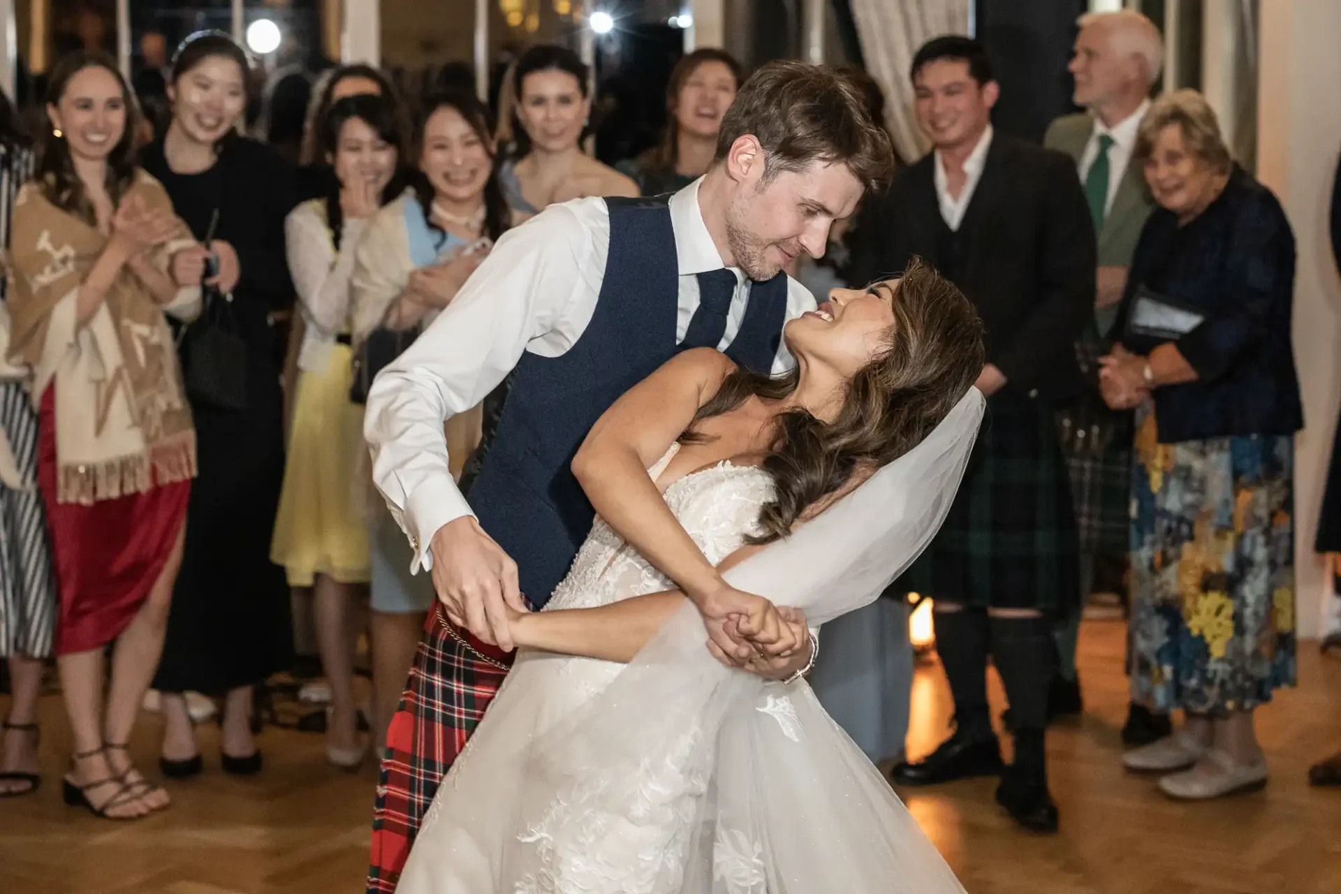 A couple in wedding attire dance joyfully, surrounded by clapping guests in a warmly lit room.