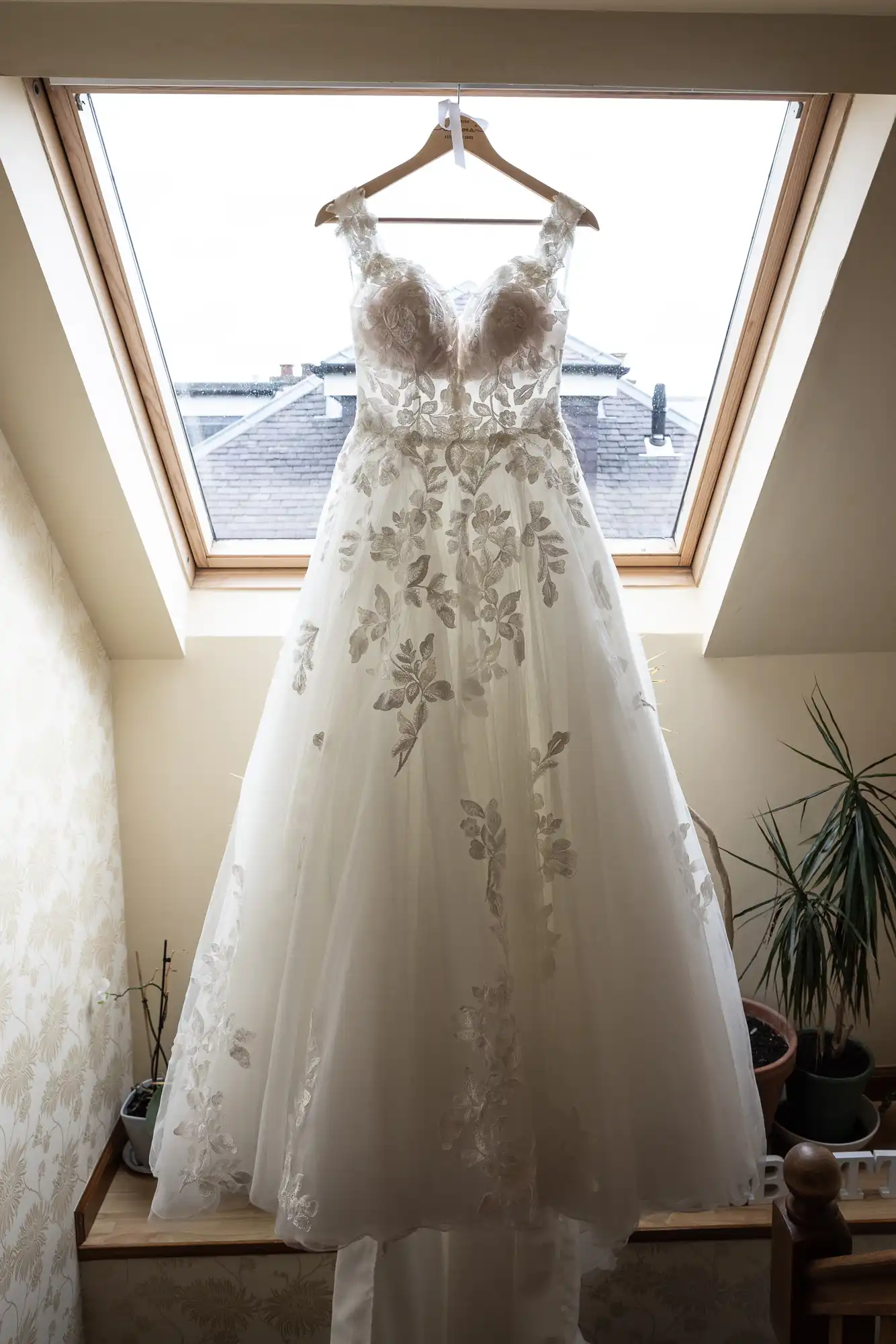 A white wedding dress with floral embroidery is hanging on a wooden hanger in front of a skylight window.