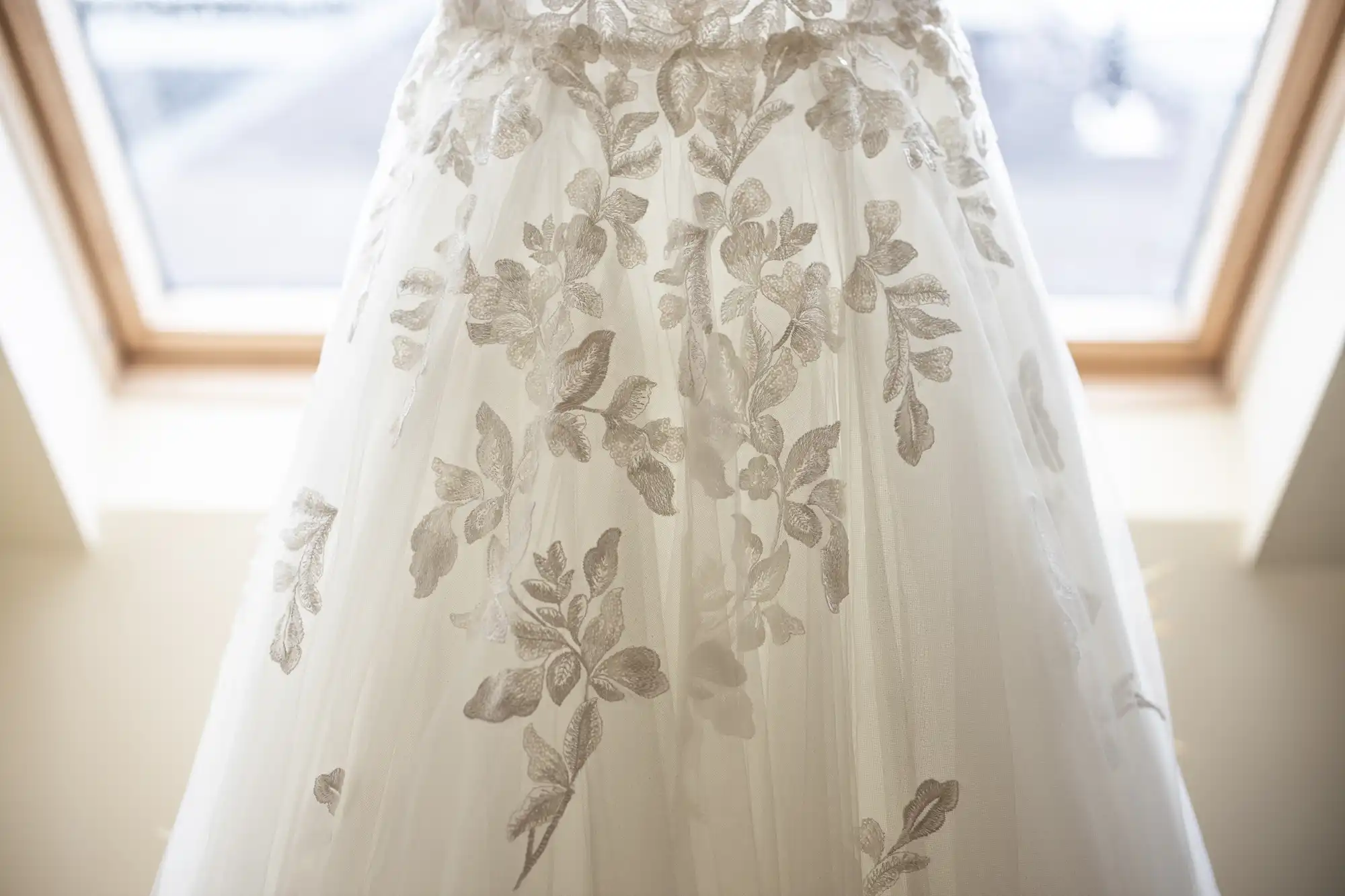 Close-up of a white wedding dress with intricate floral lace detailing, photographed against a bright window background.