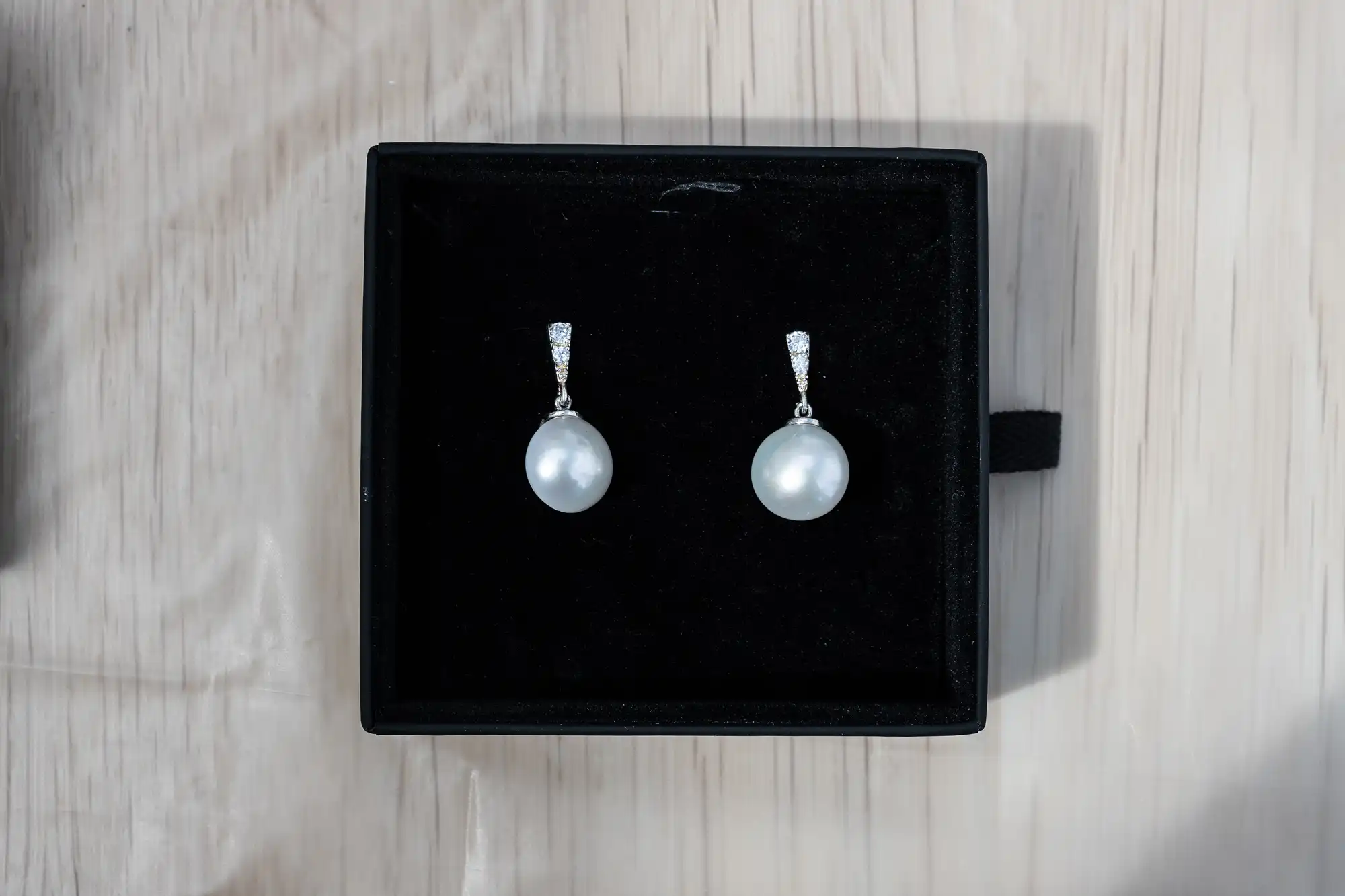 A pair of pearl drop earrings with small, clear stones set in silver, displayed on a black velvet-lined box.