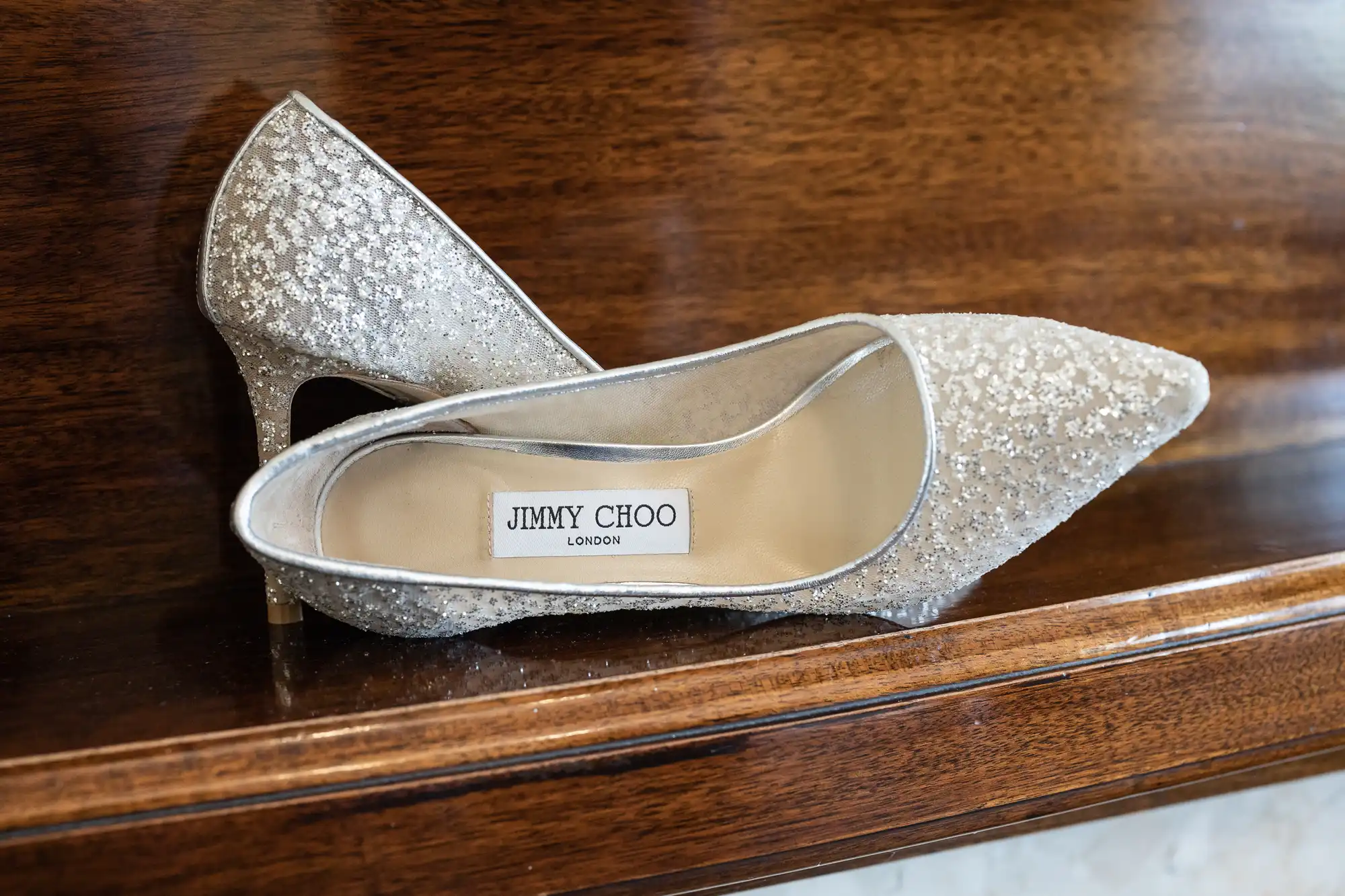 A pair of silver glitter high-heeled shoes with the label "JIMMY CHOO LONDON" on a wooden surface.