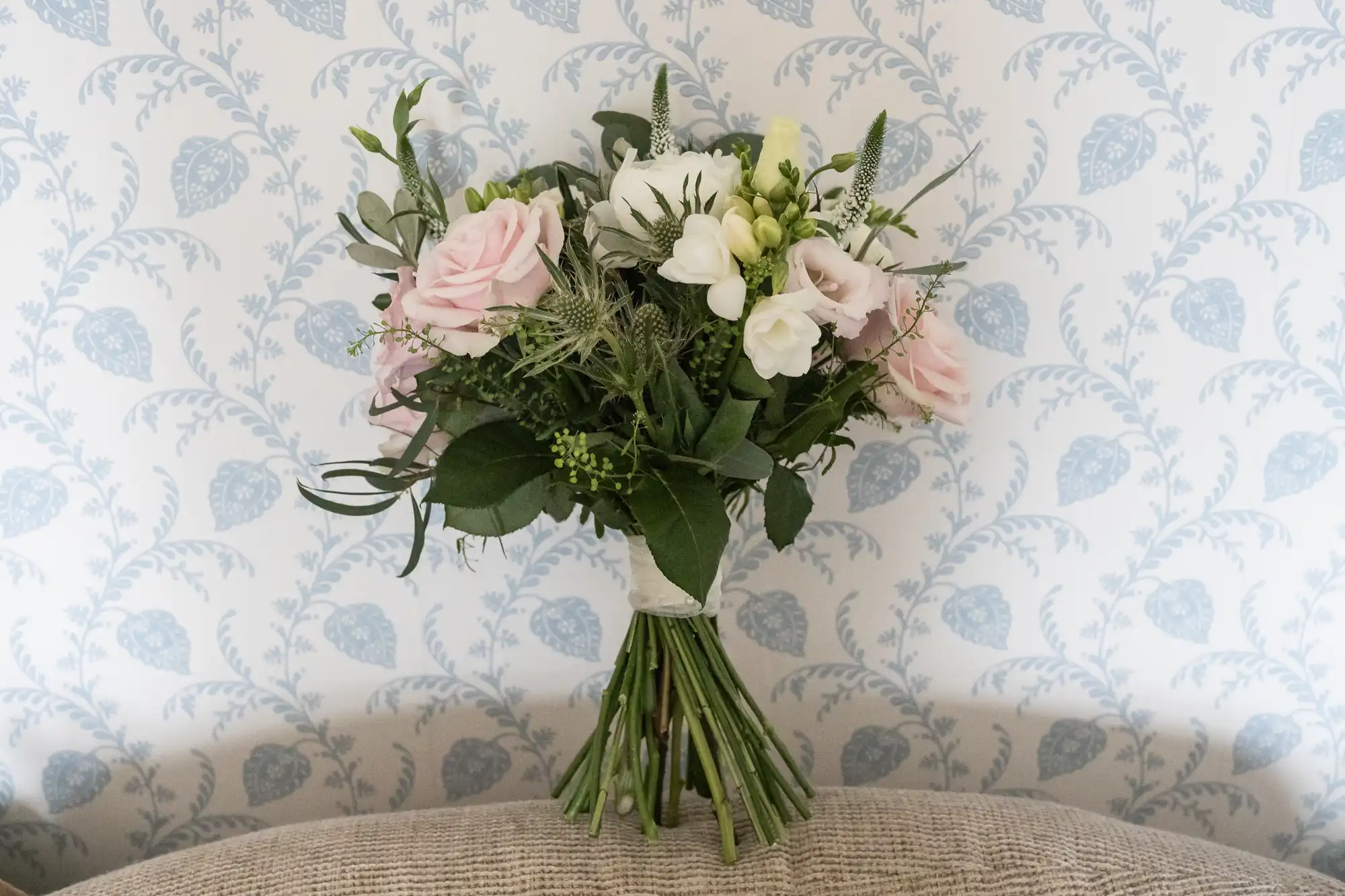 A bouquet of assorted flowers, including pink and white roses, is placed against a cushion with a light blue, floral-patterned wallpaper background.