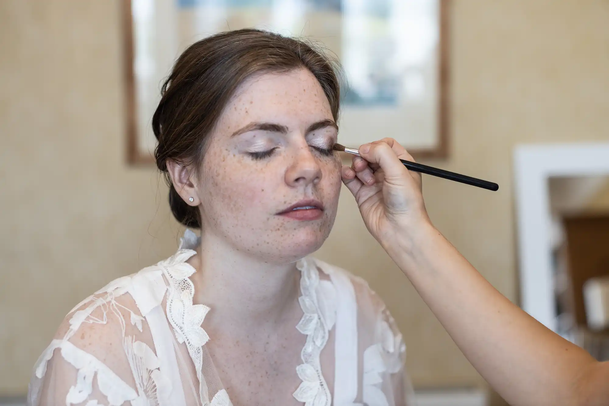 A person with closed eyes is having makeup applied with a brush by another person. The background shows a framed picture and a mirror.