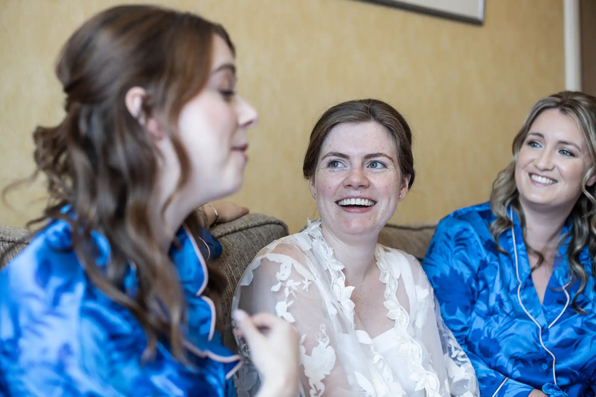 Three women in blue and white robes are sitting on a couch, engaging in a casual conversation and smiling.