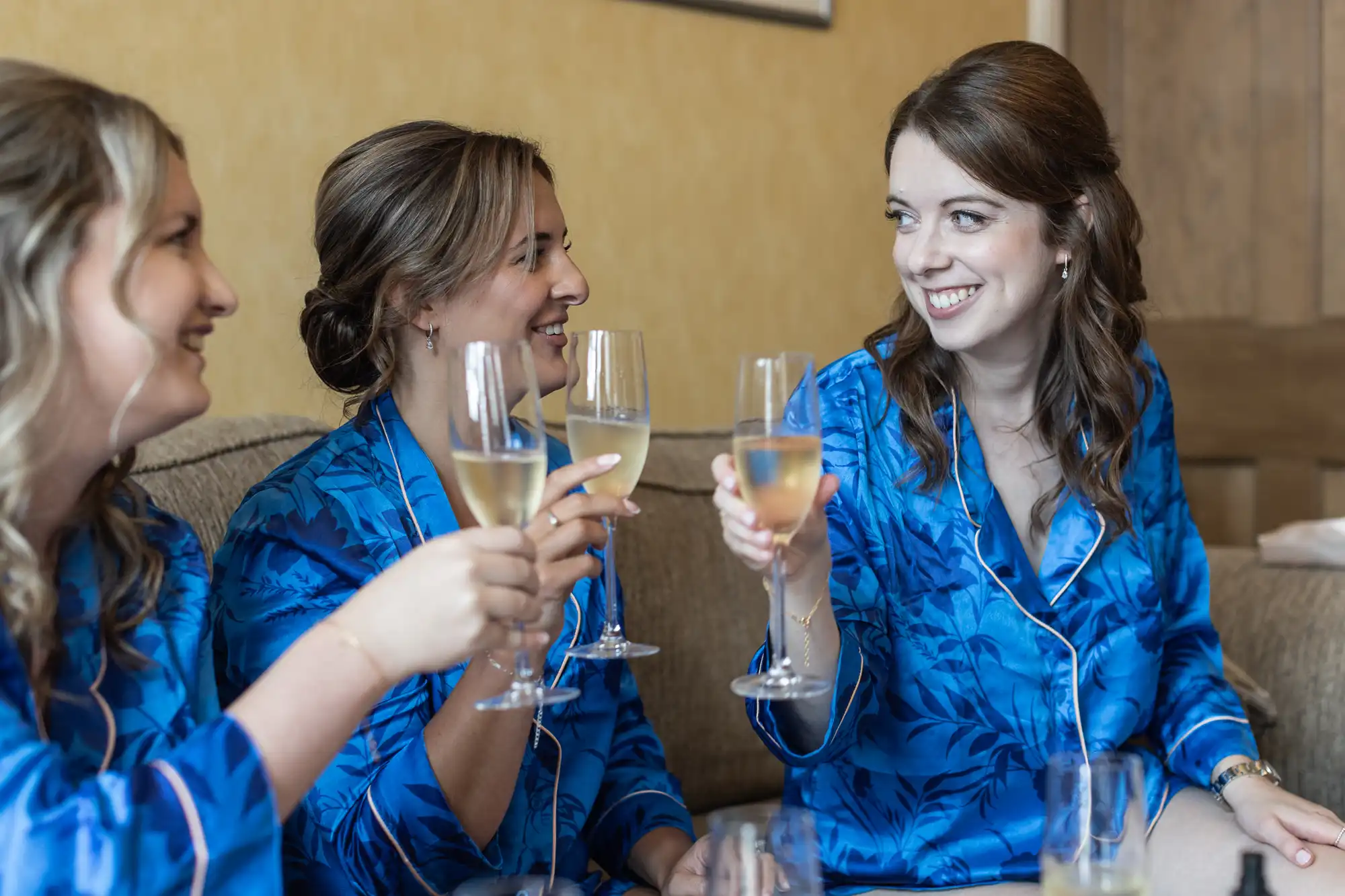 Three women wearing matching blue robes smile and raise champagne glasses while sitting on a couch.