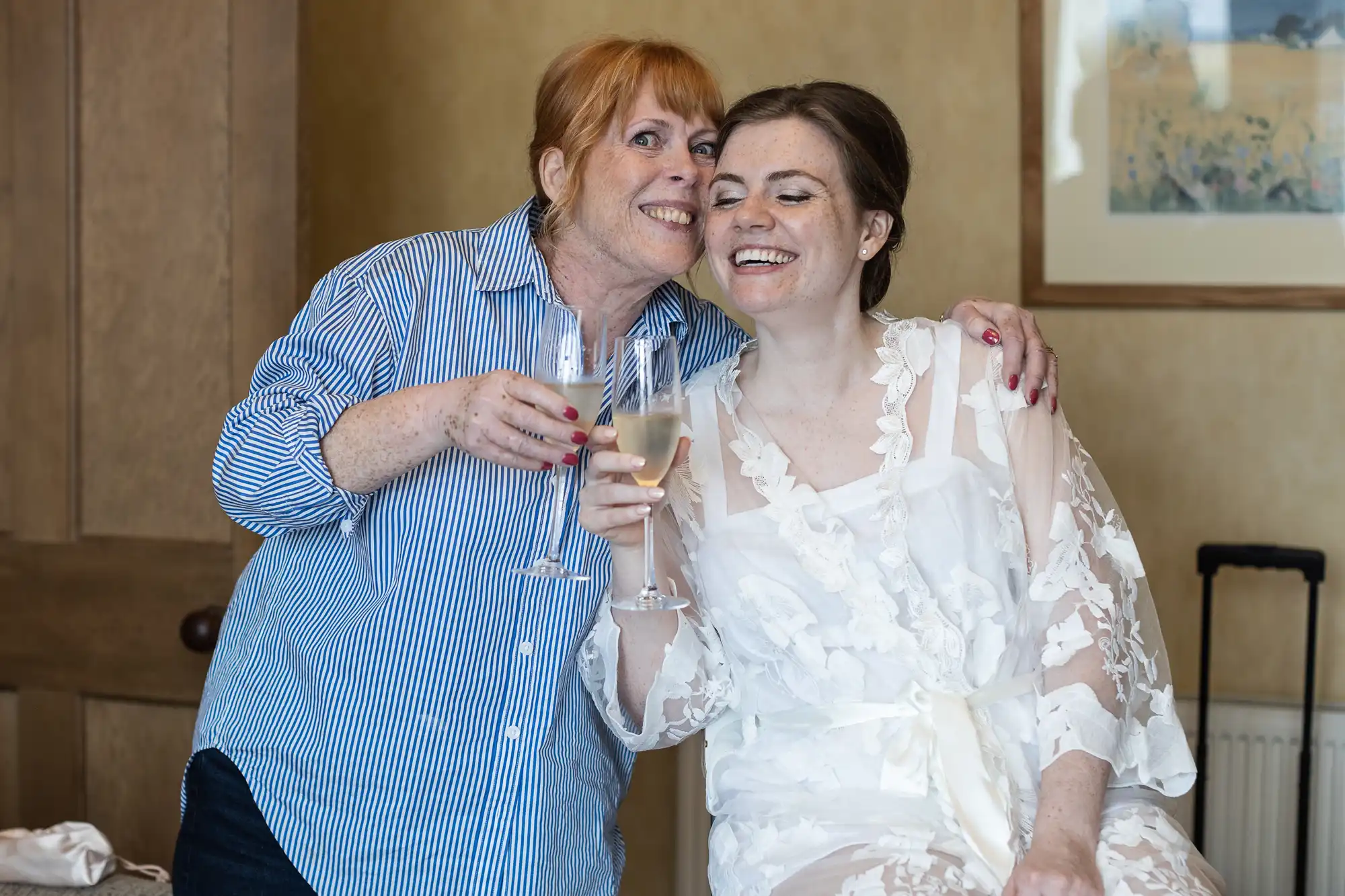 Two women smile and clink champagne glasses. One is wearing a striped shirt, and the other is in a lace white robe, possibly preparing for a wedding.